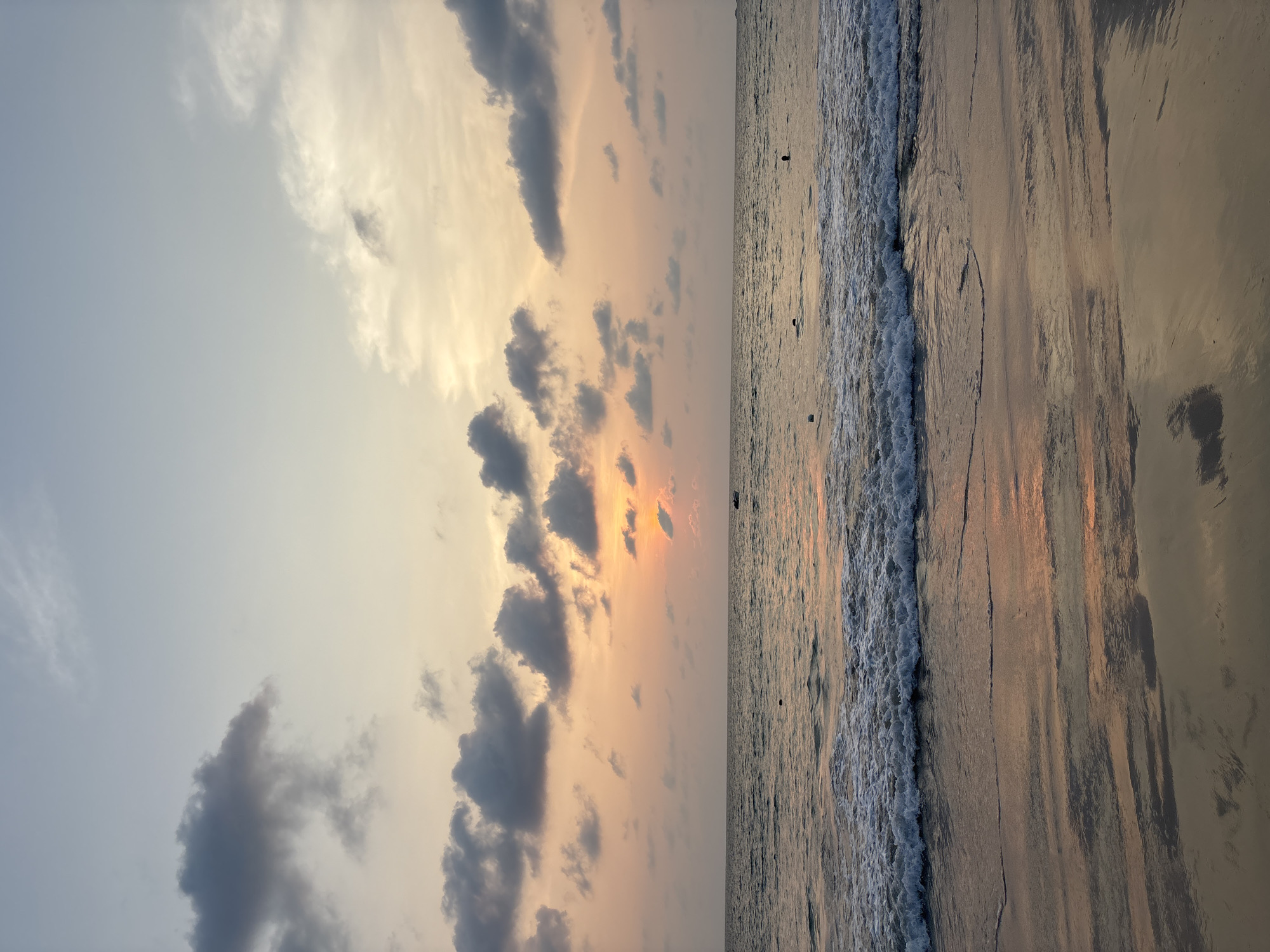 The image depicts a beach scene in Choeng Thale, Thailand at sunset. On the left side of the image is the sky with a gradient from light blue to orange-pink, filled with scattered cumulus clouds. The sun is setting behind a few dark gray clouds. On the right side is the beach. The tide is coming in, with waves lapping at the shore. The wet sand reflects the colors of the sky, creating a mirrored effect. Three silhouetted figures are in the water.