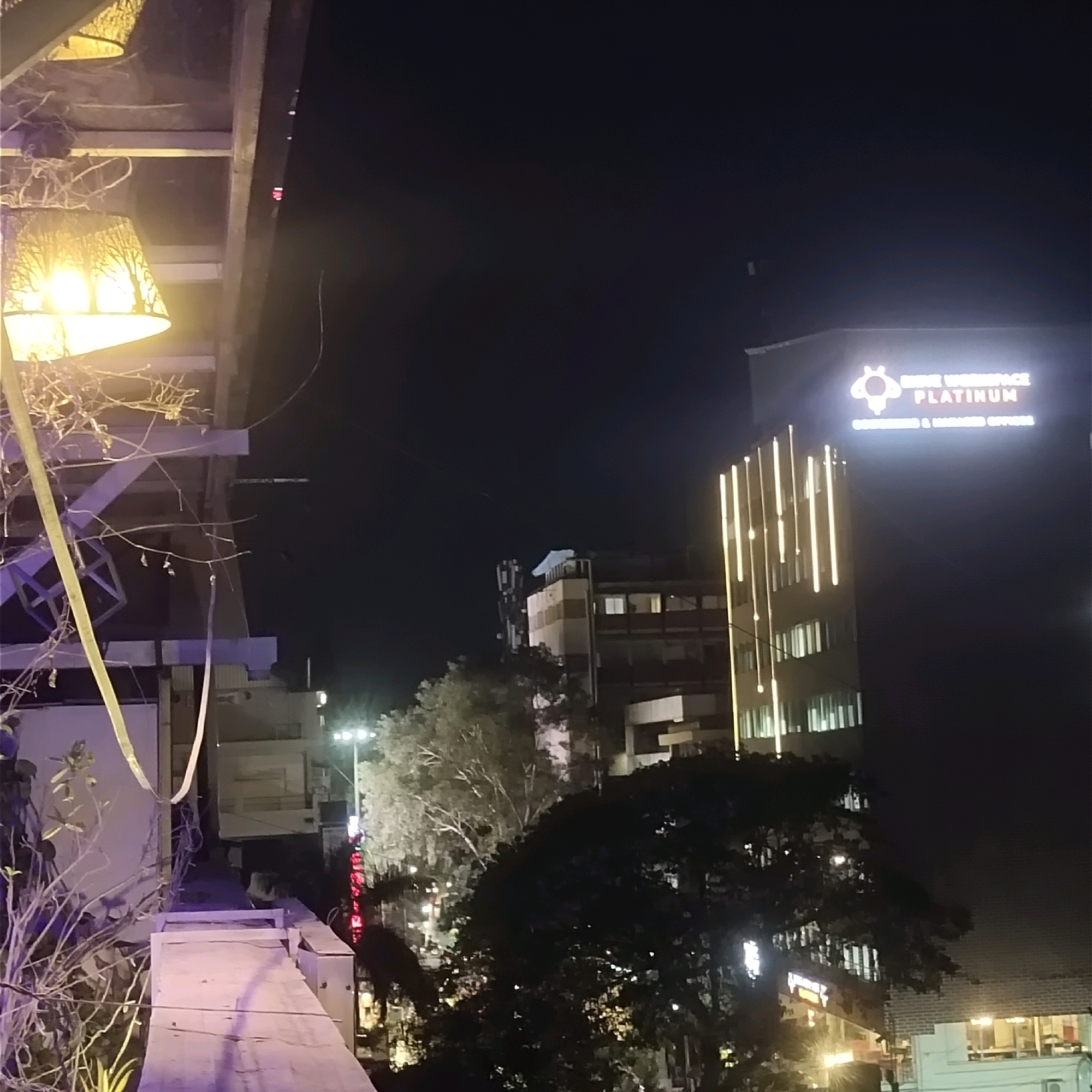 The image captures a nighttime scene in Bengaluru, India, focusing on urban architecture illuminated by artificial lights. In the foreground, a portion of a wooden structure with a hanging lamp shade is visible, casting a warm, diffused light. The lamp shade has a distinctive pattern of tree-like branches. To the right, a modern, multi-story building stands prominently, its facade adorned with vertical, yellow LED lights that create a striking visual effect. At the top of this building, a illuminated sign with a bee logo and the text "MINE WORKSPACE PLATINUM" and "SERVICED & MANAGED OFFICES" is clearly visible. Further into the background, other buildings with illuminated windows and streetlights can be observed, contributing to the bustling city ambiance. Dense foliage from trees partially obscures some of the structures, adding a natural element to the urban landscape. The sky is dark, typical of a nighttime setting in a city. No people are discernible in the image.