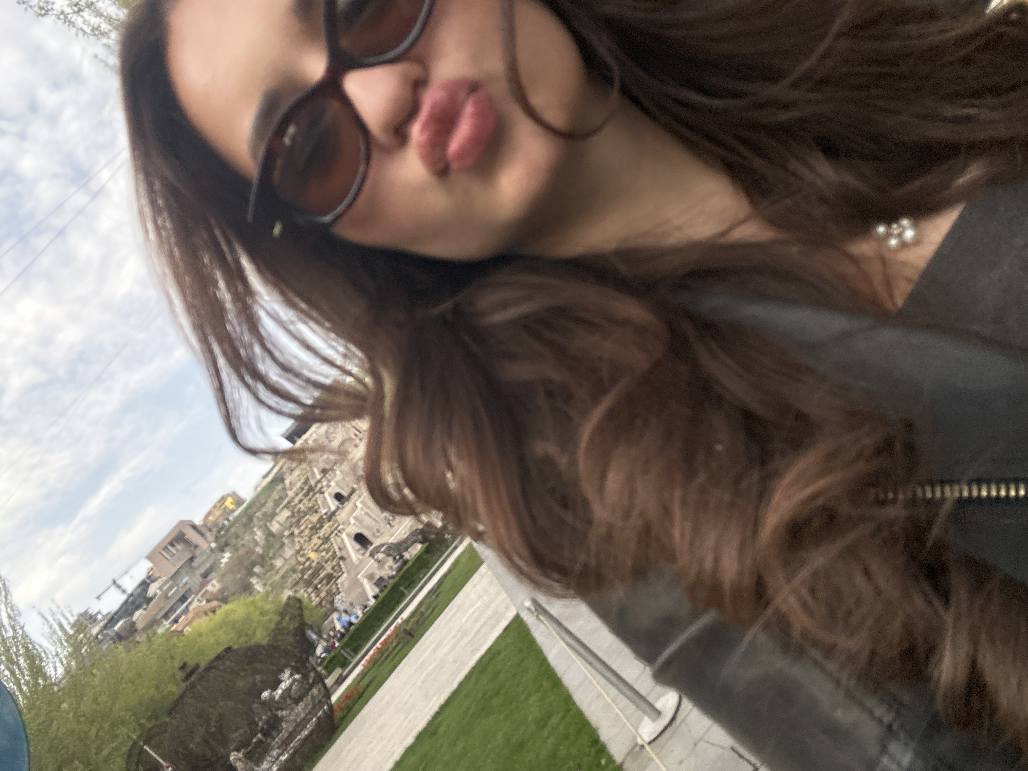 A selfie taken outdoors in Yerevan, Armenia. A woman wearing sunglasses and a dark jacket is making a kissing face toward the camera. In the background, visible over her shoulder, are trees, green lawns, walking paths, a monument, buildings, and construction cranes against a cloudy sky.