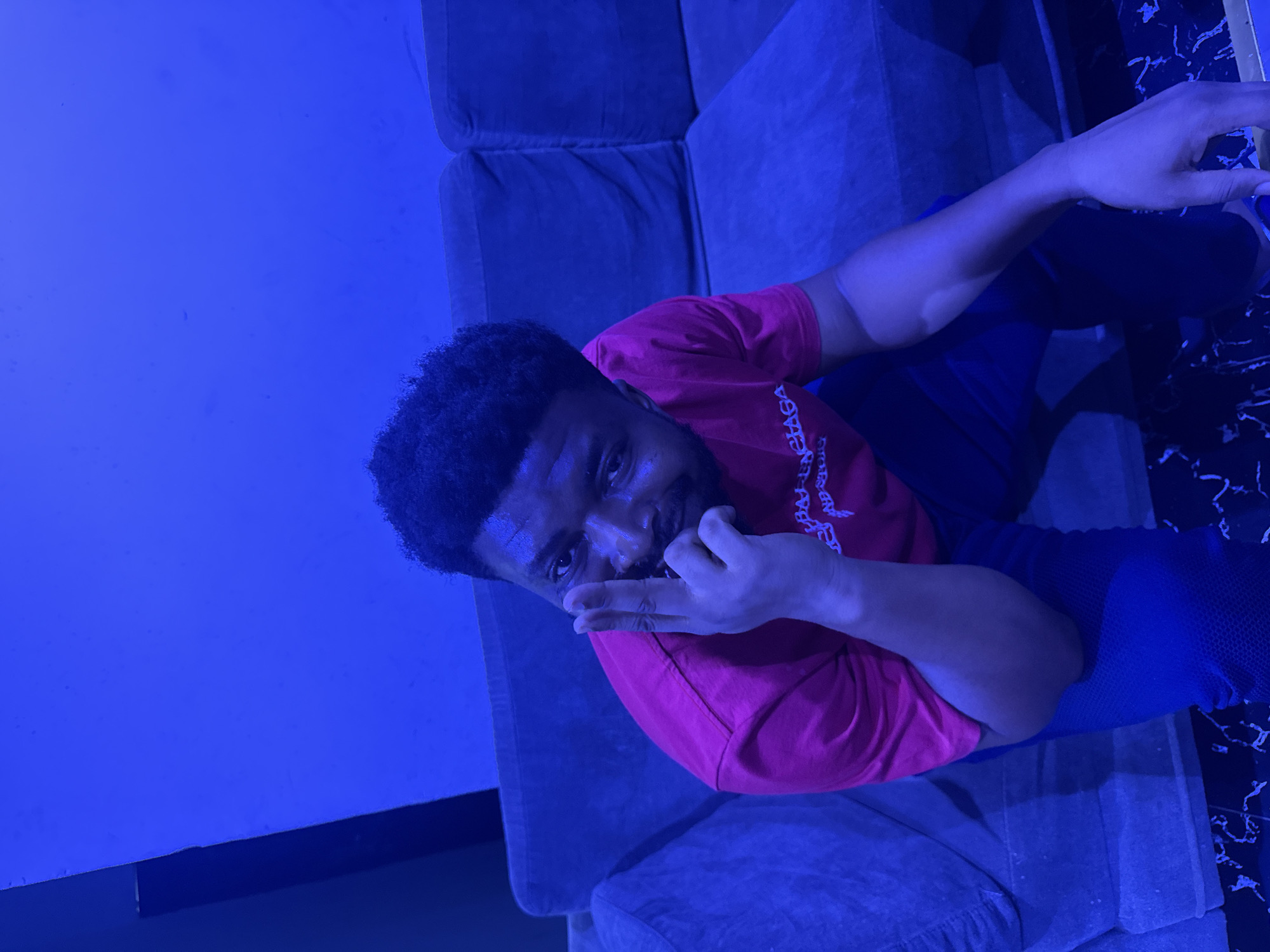 A medium-shot of a man of African descent sitting on a grey sofa. He is wearing a red T-shirt with white lettering and dark pants. His right hand is raised to his face, touching his mouth. The photo is taken in a room with blue lighting. The exact location within Abuja, Nigeria is impossible to ascertain from the image.