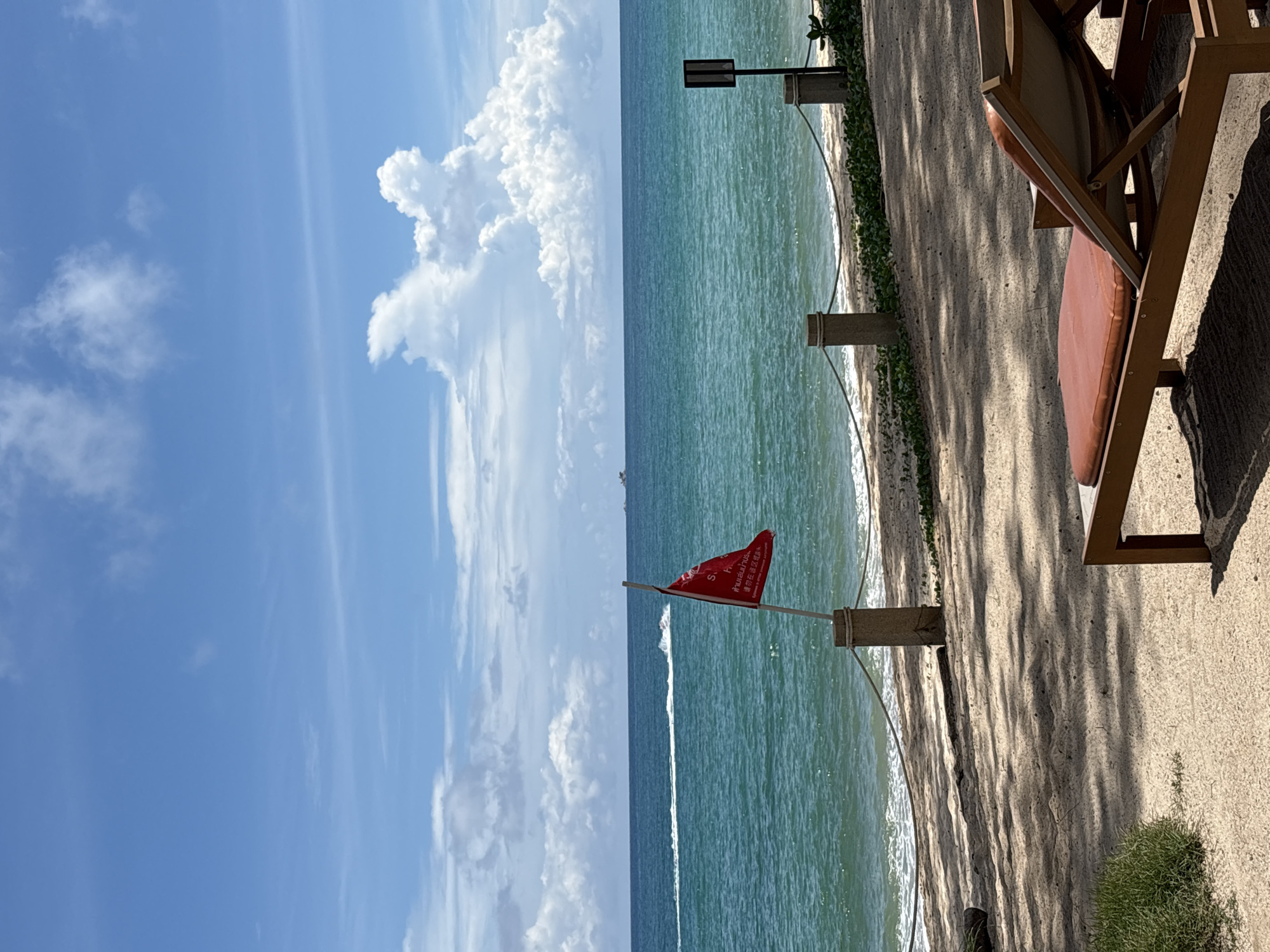 A red flag flies from a wooden pole on a sandy beach in Choeng Thale, Thailand. The flag has text in white, black, and red. The turquoise ocean stretches to the horizon, meeting a blue sky dotted with white clouds. A jet ski leaves a white wake in the distance, and a ship is visible on the far horizon. A rope barrier is strung between stone posts in front of the water. In the foreground, a beach chair is positioned on the sand, casting a shadow. Green grass grows to the left of the chair.