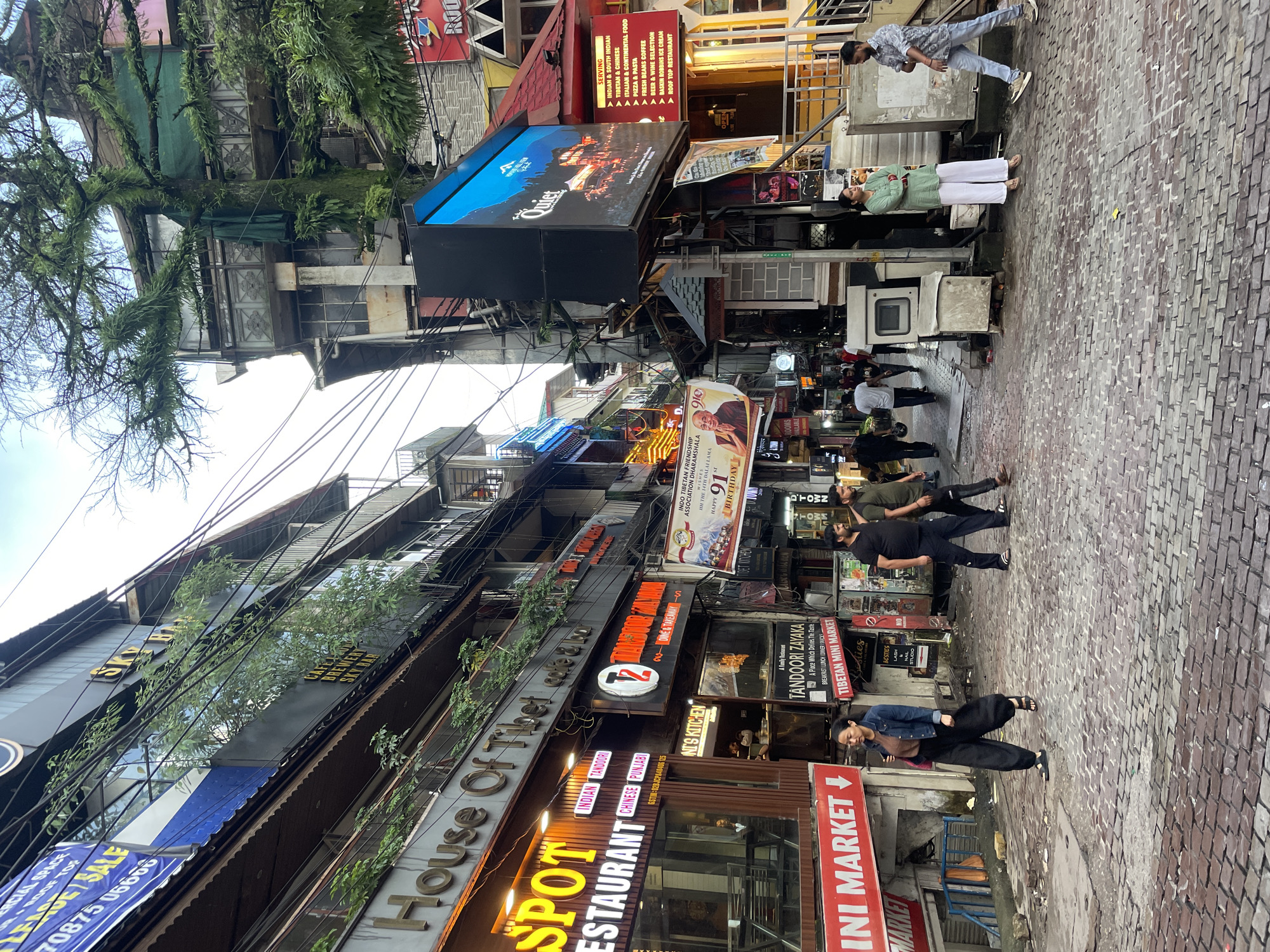 The image depicts a narrow, cobblestone street in Bhagsu, India, lined with multi-story buildings housing various shops and restaurants. The architecture features balconies, awnings, and a dense network of overhead electrical wires. On the left, a storefront with neon signage reads "SPOT RESTAURANT" and displays "INDIAN TANDOORI CHINESE PUNJABI." Above it, a sign for "House of Tibet cafe & bar" is visible. Further down the street on the right, a large digital billboard displays an advertisement for "Quiet." Below the billboard, a red sign lists "SERVING INDIAN & SOUTH INDIAN, ITALIAN & CONTINENTAL FOOD, PIZZA & PASTAS, FRESH BREEZE COFFEE, BEER & WINE SELECTION, BASKIN ROBBINS ICE CREAM, ROOFTOP RESTAURANT." Several people are walking along the street. A woman in a denim jacket and black pants is walking towards the viewer on the left. On the right, a man in a green top and white pants stands with his arms crossed, facing away from the viewer, while another man in a patterned shirt and jeans walks past him. In the background, more people are visible, and the street continues into the distance, showing more buildings and signage. The sky is overcast.