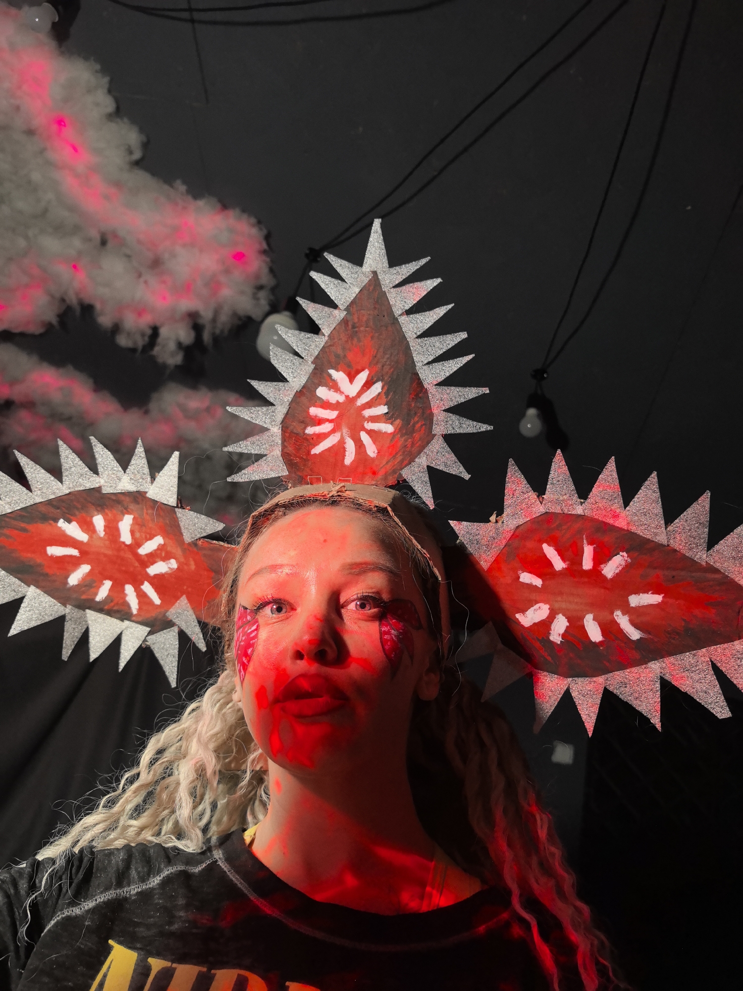 A medium close-up shot captures a person, likely a young woman, in an elaborate costume and makeup, illuminated by dramatic red lighting. She looks directly at the camera with wide, alert eyes and slightly parted lips, conveying an intense expression. Her long, curly, blonde hair cascades over her shoulders.
She wears a distinctive headpiece, possibly handmade, featuring three large, spiky, red and silver shapes resembling eyes or a monster's maw. Each shape has a red interior with white dash-like patterns and a jagged, silver-painted outline. One central piece sits atop her head, while two larger ones extend horizontally to either side.
Red makeup is visible on her cheeks and under her eyes, appearing splattered or painted in streaks. Her face, neck, and visible chest are heavily cast in red light. She wears a dark, possibly black, t-shirt with faint yellow text on the front, of which "NUD" or "ND" is partially discernible.
The background is dark and indistinct, suggesting an indoor setting. To the upper left, thick, white smoke or fog is dramatically lit with red, creating an eerie effect. Overhead, black cables and a bare lightbulb are visible against the dark ceiling. The scene suggests a themed event, party, or performance in Noviy, Russia, characterized by its dramatic lighting and costume.