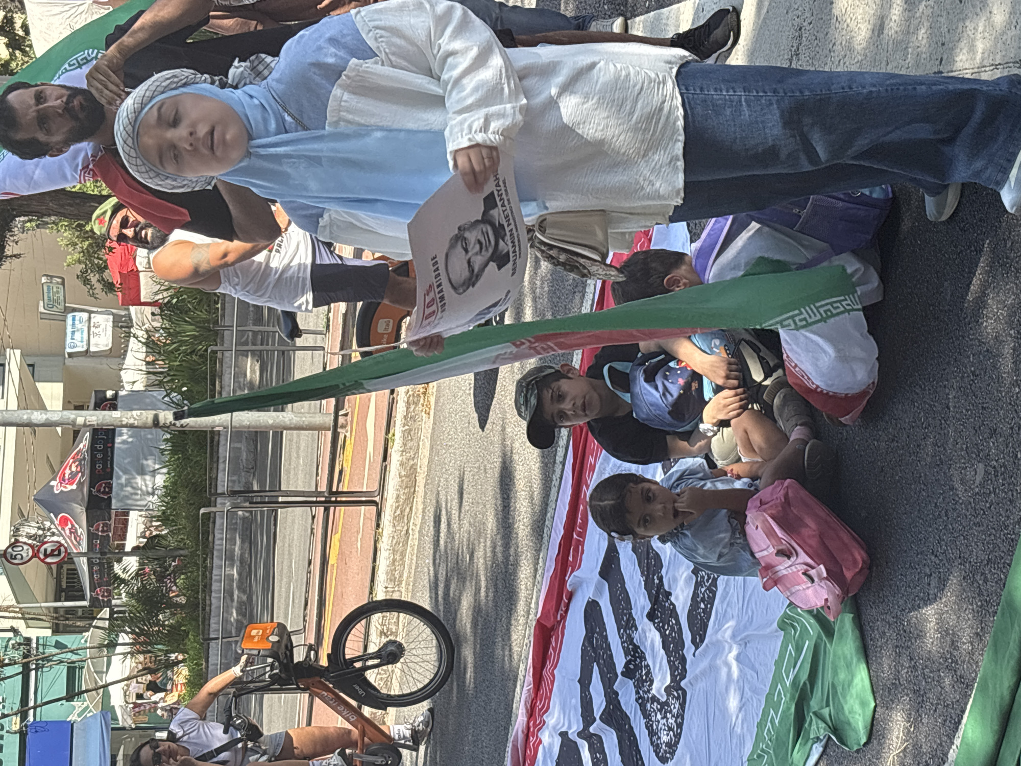 A medium shot depicts a group of individuals, including children, seemingly engaged in a protest or demonstration on a street, potentially in São Paulo, Brazil, based on the presence of signage in Portuguese.
A child in a light blue hijab is holding a sign bearing a portrait of Benjamin Netanyahu, with text in Portuguese translating to "inhumanity". Adjacent to her are other individuals, some holding a large Iranian flag. The children are seated on the street, with backpacks and personal belongings beside them.
The environment includes a bike-sharing stand with "bike itau" branding, and "Uber" indicated on one of the bikes. In the background, there is a sign with "pastel do jap" visible. Additionally, there are street signs and buildings suggesting an urban setting.