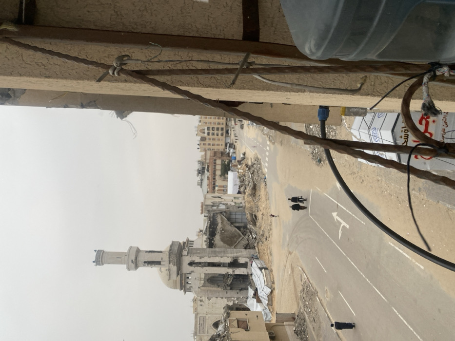 An elevated view captures a heavily damaged urban landscape in Khan Yunis, Palestinian Territory, under an overcast sky. The foreground shows a partial structure with a dark grey water tank, a metal railing, and exposed wires/conduits. Below this, a commercial sign with red Arabic script is partially visible. The midground features an asphalt road with white lane markings and a right-turn arrow, bordered by extensive areas of dirt, sand, and construction debris.
In the left-center background, the severely damaged Great Mosque of Khan Yunis is prominent, featuring a tall, slender minaret that remains largely upright despite visible damage to its base. The main mosque building exhibits significant structural collapse, with large sections of its walls destroyed and the dome partially intact but compromised. Adjacent to and surrounding the mosque are numerous buildings in various states of destruction, reduced to piles of rubble, twisted metal, and fragmented concrete walls. Towards the mid-right, a wide expanse of debris extends to a straight section of the road in the distance, where a long line of people or vehicles is visible.
On the asphalt road in the midground, three individuals, dressed in dark clothing, walk together away from the viewer. Further back on the left side of the road, a solitary individual also walks away. Another small figure is discernible in the debris-filled area between the road and the mosque.