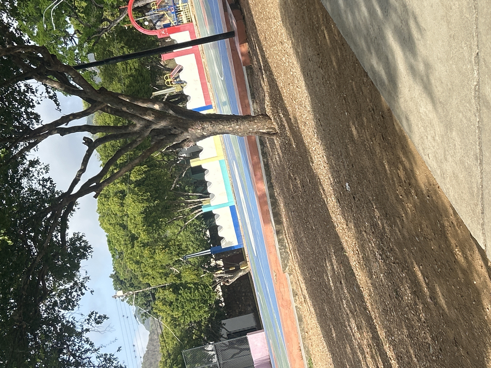 The image depicts an outdoor recreational area, possibly a park or schoolyard, in Santa Ana, Venezuela. A vibrant, multi-colored court, likely for basketball or a similar sport, is visible with blue, yellow, and red markings. The court is bordered by a low wall featuring a decorative pattern of white, blue, and yellow sections. Adjacent to the court, a playground with red and yellow slides and climbing structures is partially visible. A large tree with a thick trunk and spreading branches dominates the foreground, casting dappled shadows on the ground, which is covered in light brown gravel and dirt. In the background, lush green trees and hills under a partly cloudy sky suggest a tropical or subtropical environment. A utility pole with wires can be seen amongst the foliage. The overall impression is of a well-maintained public space.