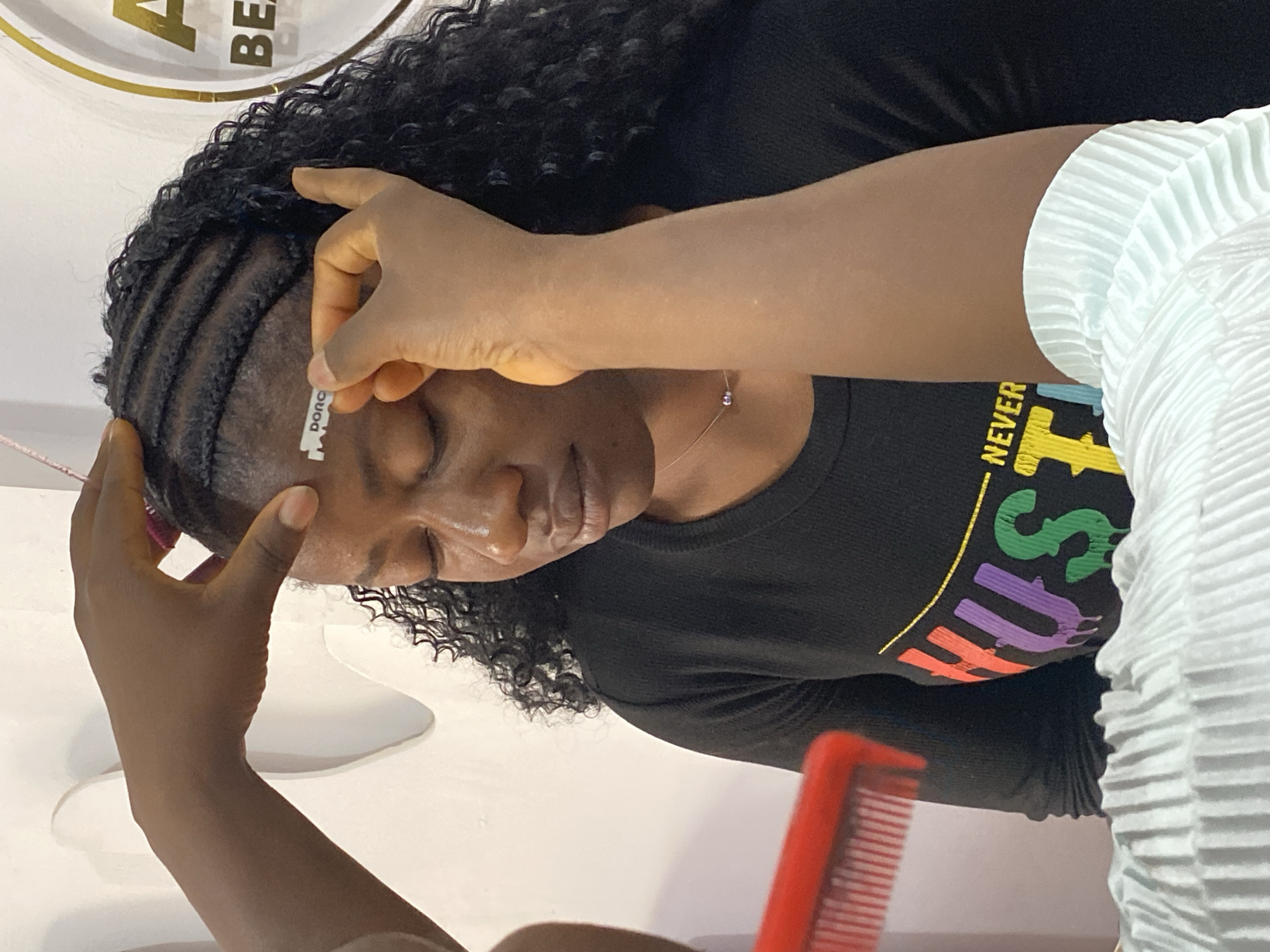 This vertically oriented close-up image captures a hair grooming session likely taking place in a salon in Bukuru, Nigeria. The primary subject is a person with dark, curly hair, whose scalp features neatly braided cornrows. Their eyes are closed, indicating focus or relaxation during the process. They are wearing a black t-shirt adorned with colorful, partially visible text, including the word "NEVER" and other letters like "HUTS." A delicate silver necklace is also visible around their neck.
A second person's hands are actively involved in styling the subject's hair. One hand carefully holds a small, rectangular razor blade, clearly branded "DORCO," near the subject's temple, suggesting a precise hairline clean-up or definition. Another hand supports the forehead area. In the lower foreground, a red comb with widely spaced teeth is partially visible.
The indoor setting features a white background wall with a gold circular design and partial text that reads "BEA," likely part of a salon or beauty establishment's signage. A light-colored, possibly pleated garment worn by the stylist is visible on the right side of the frame.