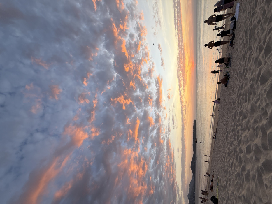The image displays a sunset scene on a wide sandy beach in Choeng Thale, Thailand. The upper two-thirds of the frame are dominated by a sky filled with scattered clouds, exhibiting prominent orange and pink coloration from the setting sun, contrasting with areas of grey and light blue. A small, intense orange-red sun disk is partially visible on the horizon, slightly right of the central vertical axis, casting a glow over the lower sky and water. A single dark bird is visible in flight in the upper right quadrant.
The middle ground is occupied by a calm body of water, likely the Andaman Sea, reflecting the sky's colors. Several boats are present in the distance; one larger vessel is situated left of center, and multiple smaller crafts are scattered further out. A line of dark, vegetated hills forms the distant background along the horizon, with a headland visible on the far right.
The foreground consists of light-colored sand, showing texture from ripples and footprints. Approximately 25-30 individuals are present on the beach or wading in the shallow water. On the far left, several people are lying prone or reclining on towels or mats. Towards the mid-right, a group of five individuals, predominantly male, are walking from left to right, silhouetted against the water. Other individuals are standing, sitting, or wading near the water's edge. A dark bag or backpack is visible on the sand in the far left foreground, and other small items, possibly clothing or towels, are near some seated individuals.
