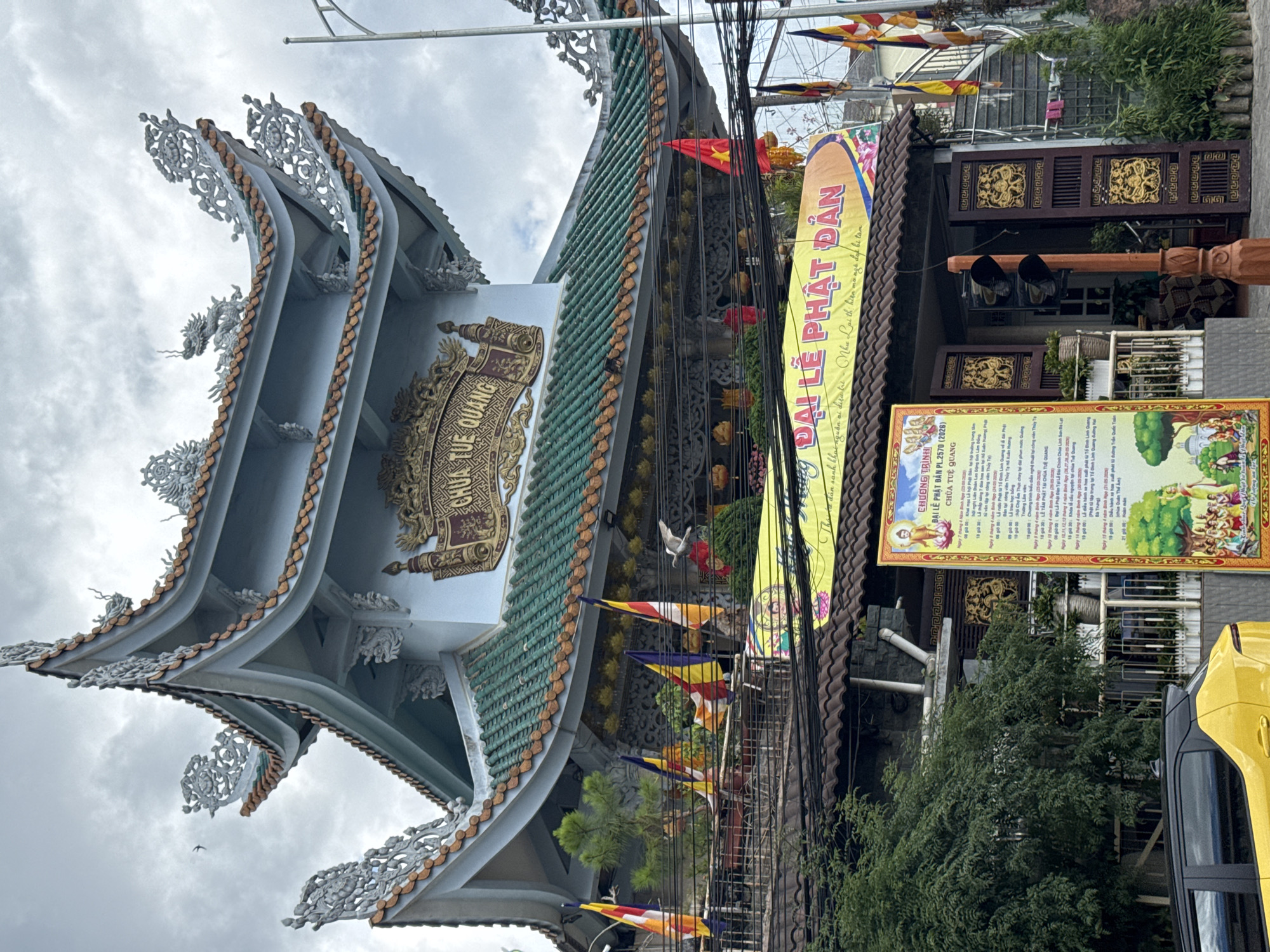 The image shows a Buddhist temple in Cam Ly - Đà Lạt, Vietnam. The temple has a multi-tiered roof with intricate carvings and decorations. There is a sign with Vietnamese writing on it indicating a Buddhist festival. Flags are displayed outside the temple. Power lines are visible overhead. There is foliage and a car in the foreground.