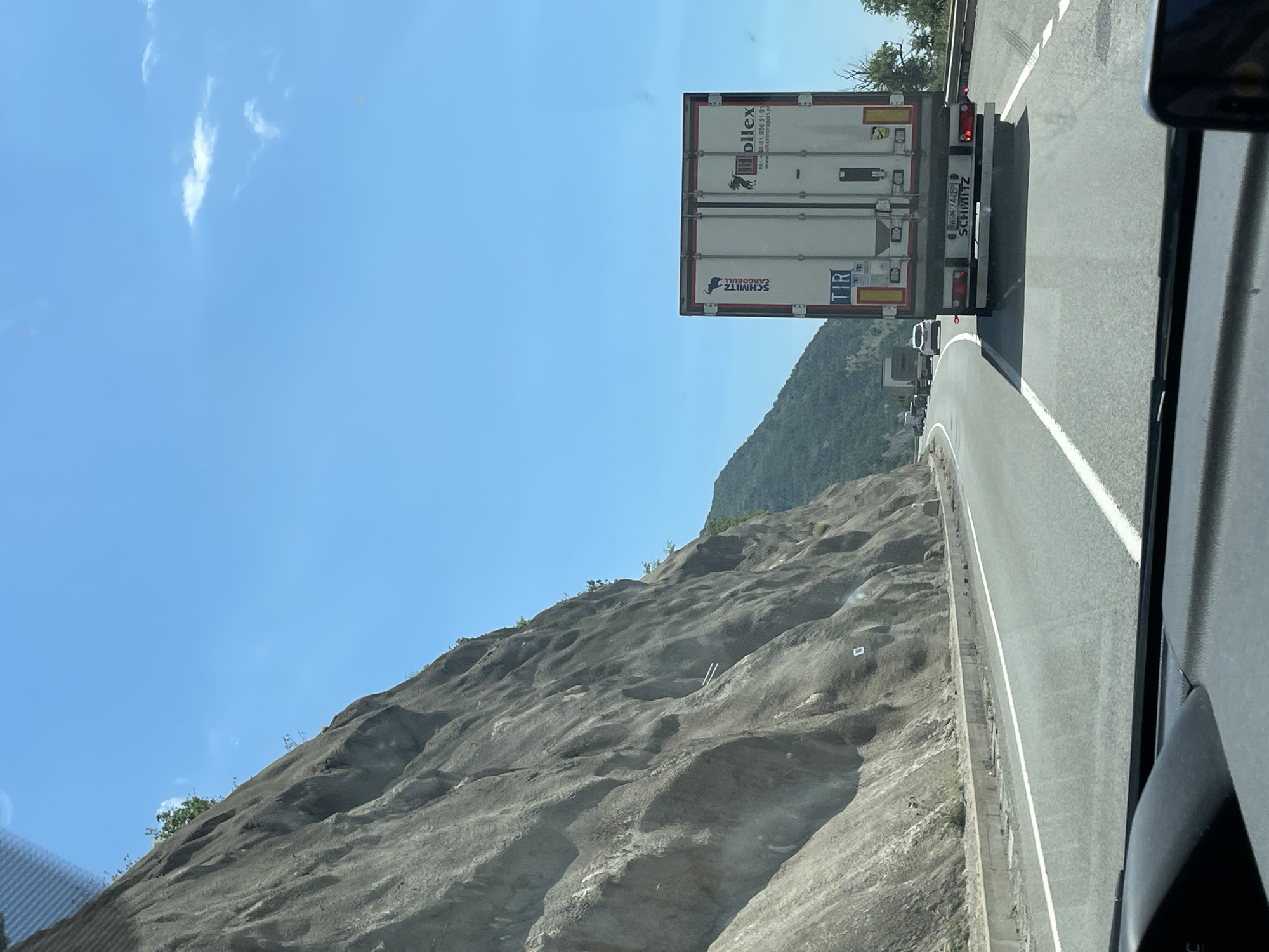 The image depicts a daytime scene from the interior of a vehicle, looking forward through the windshield onto a multi-lane highway. In the immediate foreground, a dark dashboard is visible, with reflections appearing on the windshield above.
Directly ahead, a white semi-trailer truck occupies the lane. The rear of its trailer features "SCHMITZ CARGOBULL" text vertically on the left door and a "Hollex" logo with a bull, along with a Polish phone number (+48 31 326 51 91) and website (www.hollextransport.pl), on the right door. A "TIR" plate is affixed to the left door. The truck's license plate, partially visible, displays "WSN 74492". Red taillights are illuminated.
To the left of the highway, a massive, steep rock face, grey and heavily textured, rises dramatically alongside the road. The rock exhibits vertical striations and varying contours, with small bushes and trees clinging to its surface. Visible on the rock face are multiple white markers or caps, along with what appear to be two parallel metal rods, indicating geological stabilization or monitoring. A concrete barrier separates the road from the base of the rock.
To the right, beyond the road and other vehicles in the distance, a distant, green-clad hillside is visible under a clear, bright blue sky with a few wispy white clouds. Traffic ahead appears moderate, with several vehicles visible further down the road, including another white truck and a white passenger car.
The distinctive geological features, particularly the close proximity of the road to a towering rock face, are characteristic of the DN6/E70 highway as it traverses the Iron Gates gorge near Drobeta-Turnu Severin, Romania, where the road runs alongside the Danube River through mountainous terrain.