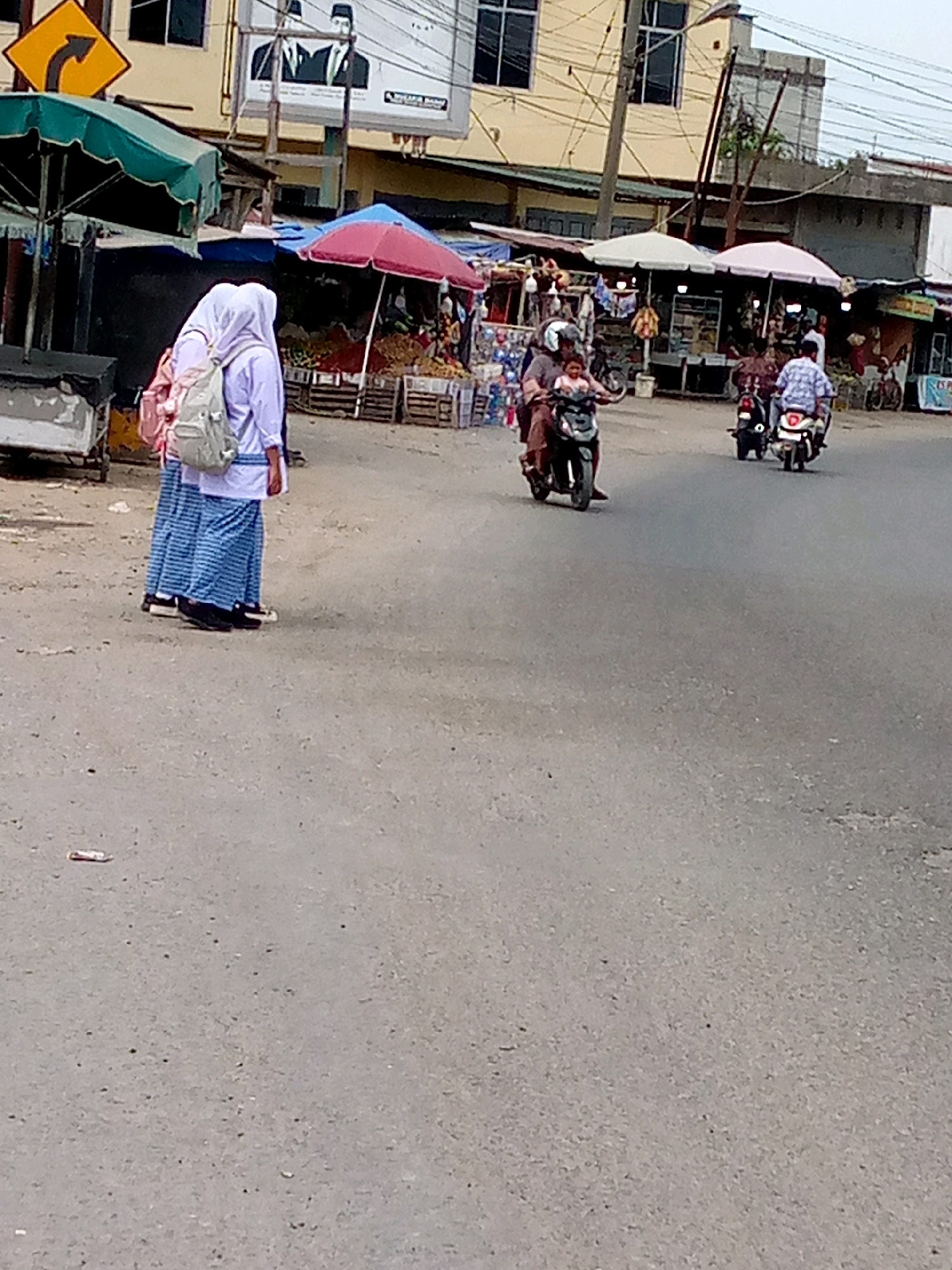 This is a street-level, medium-angle, eye-level shot of a street scene in Keudegeudong, Indonesia. In the foreground, two individuals wearing white shirts and blue and white striped sarongs are walking away from the camera. They both have backpacks on, one of which is light grey and the other is pink. To the left of them, a green market stall canopy is visible. In the middle ground, three motorcycles are present. One motorcycle on the left has two occupants, a driver and a passenger, who is a child. A second motorcycle is stopped in the middle, with its rider facing away from the camera. A third motorcycle is on the right, also with its rider facing away. In the background, there are several market stalls with umbrellas and awnings, displaying various goods. Buildings line the street, some with visible signage and advertisements. A traffic sign is also visible on the left. The pavement is asphalt.