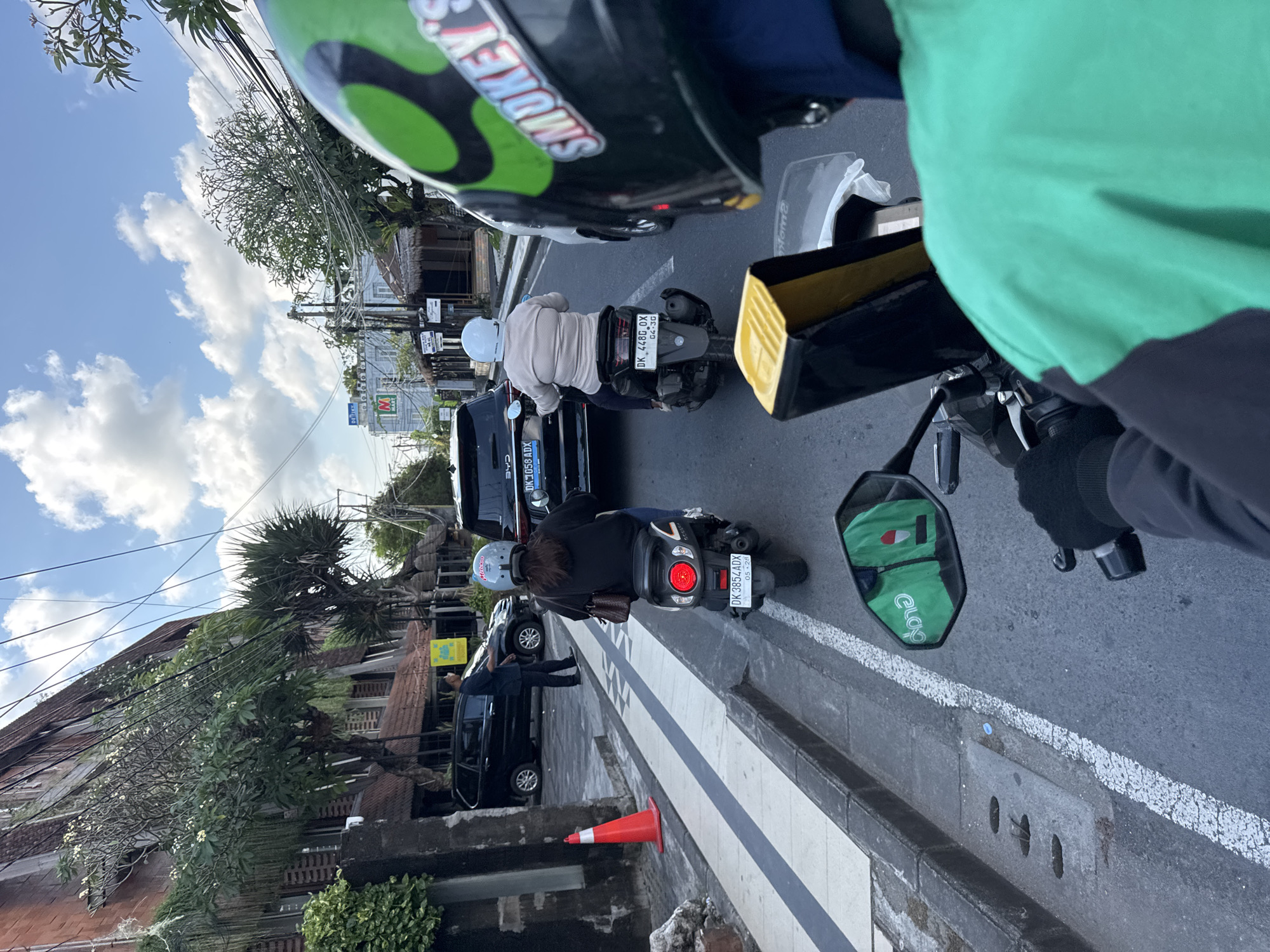 This eye-level, first-person perspective photograph captures a busy street scene in Kuta, Indonesia, from what appears to be a motorbike. The sky is bright with scattered white clouds, indicating daytime.
The foreground is dominated by parts of the motorbike from which the photo is taken, including the rider's green top, a black long-sleeved garment, and a helmet with "SMOKEY" written on it. The motorbike's mirror reflects a green bag with a logo that appears to read "eLogo" or similar, suggesting a delivery service.
In front, traffic is visible, consisting primarily of motorbikes and cars. One motorbike rider directly ahead wears a light-colored, possibly striped, long-sleeved shirt and a white helmet. Another motorbike further down the road has a rider in a black top and a grey helmet. This motorbike has a red taillight and a small brown bag hanging from it. A black car with "CHE" and license plate "DK1058 ADX" is visible, as is another car and a person standing next to it. License plates "DK 4480 OX" (with "04-30") and "DK 3854 ADX" (with "05-28") are also clearly visible on motorbikes, indicating the Bali region of Indonesia.
The road features white solid and dashed lines. A red traffic cone is positioned on the edge of the road, next to a sidewalk. In the background, buildings with tiled roofs and trees line the street. Overhead, numerous power lines crisscross the sky. A distant building shows a red "M" logo, possibly for a convenience store like Indomaret, and the text "Home" is partially visible. The scene depicts typical urban traffic and infrastructure in Southeast Asia.