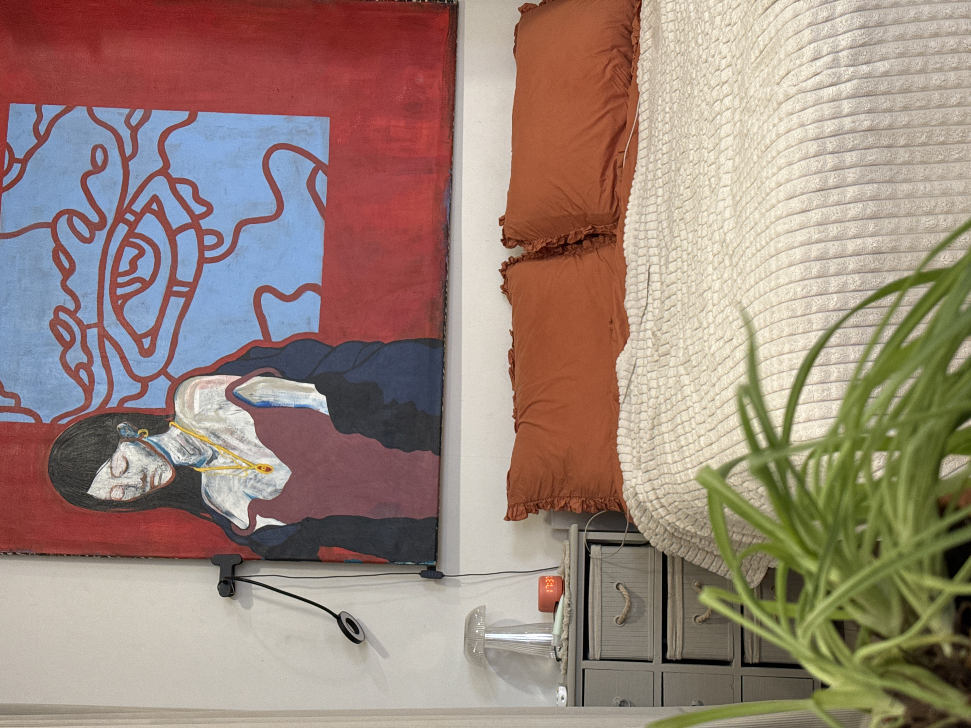 The image shows a section of an interior space with a large painting, decorative pillows, a textured blanket, a small dresser, and a houseplant. The painting depicts a figure lying down, with red and blue abstract shapes in the background. Two rust-colored pillows with ruffled edges are positioned near a white, textured blanket. A gray dresser with multiple drawers and rope handles is beneath the pillows and blanket. The bottom half of a houseplant is partially visible in the foreground, obscuring some of the scene. A lamp is affixed to the wall, positioned under the painting. An elongated glass lamp rests on top of the dresser. The location of Krasnoyarsk, Russia, cannot be determined from the provided image.