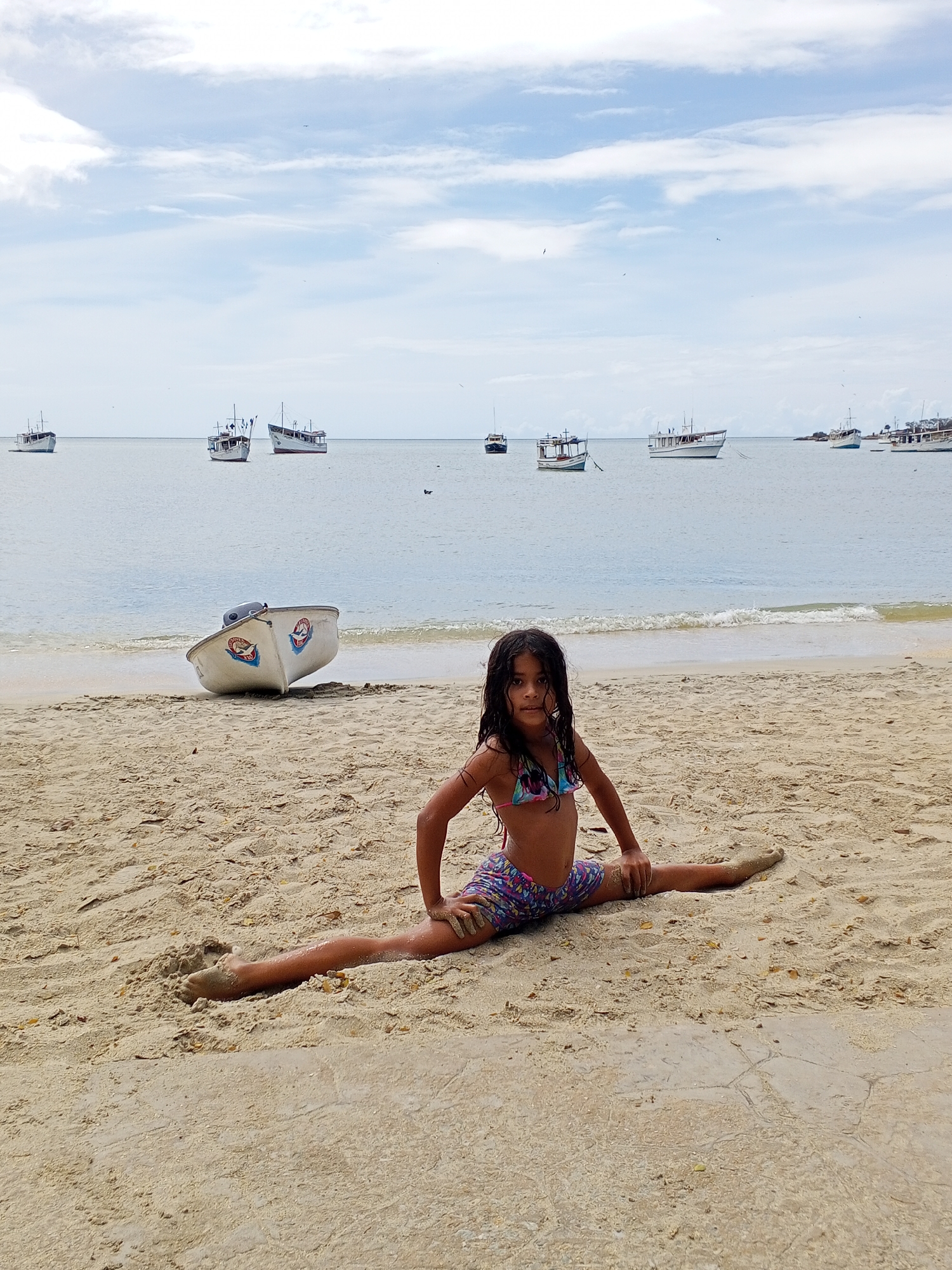 A young girl in a patterned swimsuit is performing a split on a sandy beach in the city of Unknown, Unknown. She is positioned in the foreground, looking directly at the camera. Behind her, there is a white boat resting on the sand near the water's edge. The ocean is visible in the background, with several boats floating on the water under a cloudy sky.