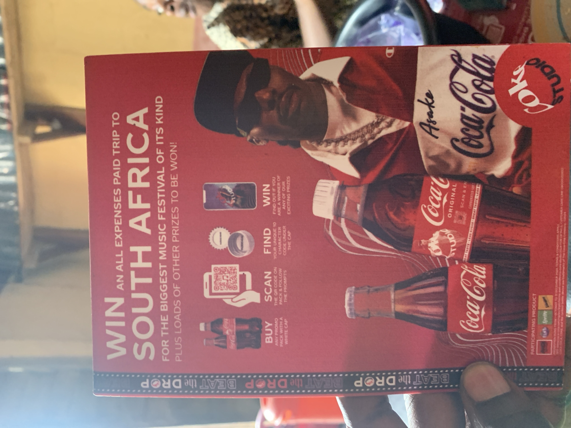 The image shows a red Coca-Cola advertisement with the text "WIN AN ALL EXPENSES PAID TRIP TO SOUTH AFRICA FOR THE BIGGEST MUSIC FESTIVAL OF ITS KIND PLUS LOADS OF OTHER PRIZES TO BE WON!" The ad features an image of Asake, the Coca-Cola logo, and instructions on how to participate in the promotion: buy a promo pack with a white cap, scan the QR code, find the unique code under the cap, and win prizes. The image appears to be taken in a residential setting, based on the background of the photo. The ad is being held by a person with dark skin. The location of Rafin Par, Nigeria is only provided to assist with the location, but is not visible in the image.