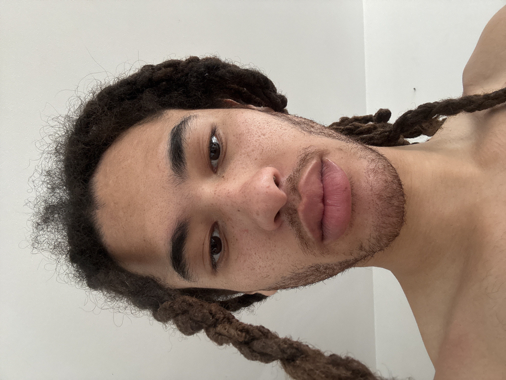 A close-up, slightly rotated shot shows a light-skinned individual with dreadlocks. The individual's face, marked with freckles, has dark eyebrows and eyes. A short, sparse beard frames the lower portion of the face, highlighting full lips. The background is a plain, light-colored wall. The location is not apparent from the image. No specific objects or actions are visible other than the presentation of the individual's face.