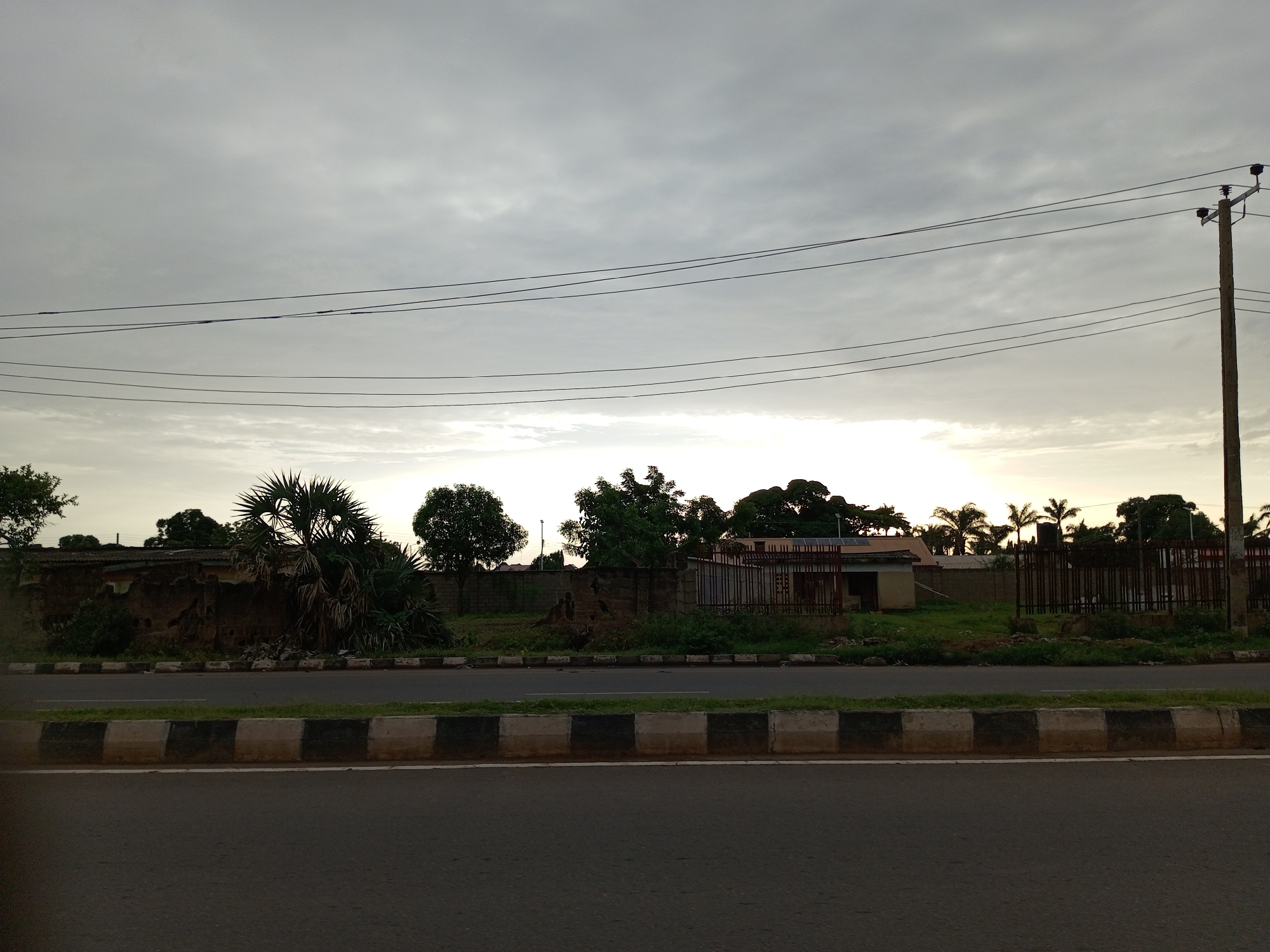 This is a medium-distance, eye-level shot of a street scene in Barden Yana, Nigeria. The foreground consists of a grey asphalt road with a black and white striped concrete barrier running along its edge, separating it from a grassy area. Beyond the grass, there is a low, crumbling brick wall, and further back, a fence with metal bars. Behind the fence, several structures are visible, including what appears to be a small building with a tiled roof and solar panels, and a water tank. Scattered trees and palm trees are present throughout the background. Overhead, multiple dark power lines stretch horizontally across the frame, supported by a wooden utility pole on the right. The sky is overcast with a bright, hazy glow towards the horizon, suggesting either sunrise or sunset. No people are visible in the image.