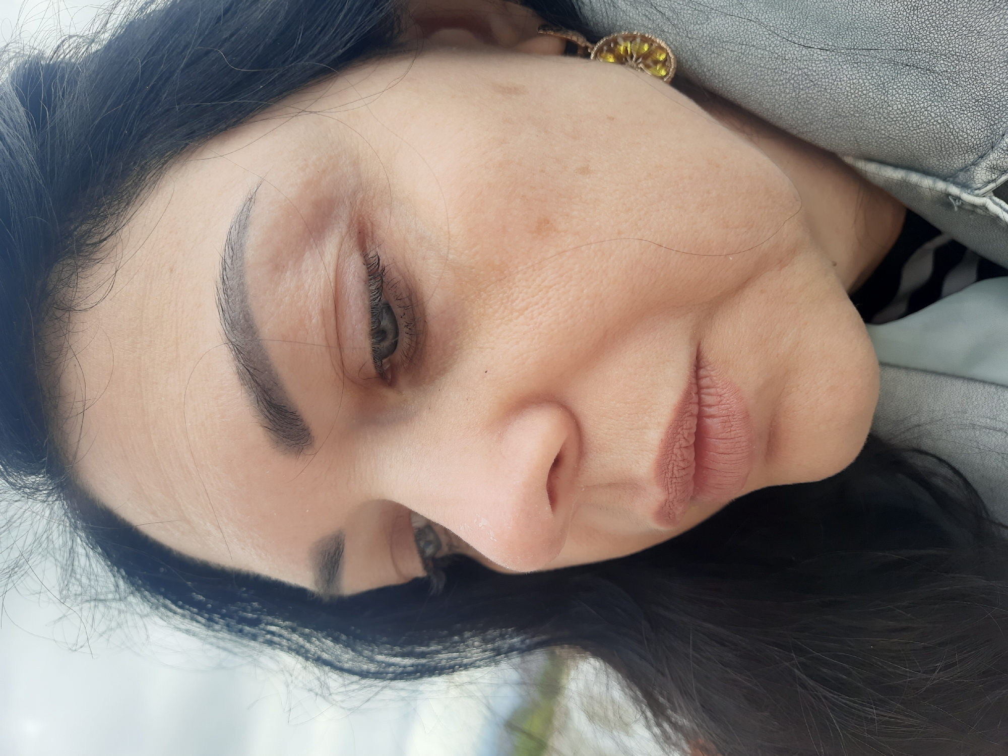 The image, set in Kumluca, Turkey, features a close-up, angled view of a woman's face and upper torso. Her head is significantly tilted, presenting a pensive or resting expression with her eyes partially closed or looking downwards. She has long, dark hair, with strands visible around her face and shoulders. Her skin is fair, showing scattered freckles and sunspots, particularly on her forehead and cheek. She has well-defined, dark eyebrows and dark eyelashes. Her prominent nose exhibits some dryness or flaking skin, as do her slightly parted lips, which are colored with a matte, light pink or nude shade. She wears a light gray or silver textured jacket or garment, with a black and white striped top visible beneath. A golden earring, featuring a clustered yellow detail, is visible near her right ear. The background is softly blurred, suggesting a brightly lit outdoor or indoor setting with indistinct elements resembling a light sky or wall and possibly some distant greenery. No text is visible in the image.