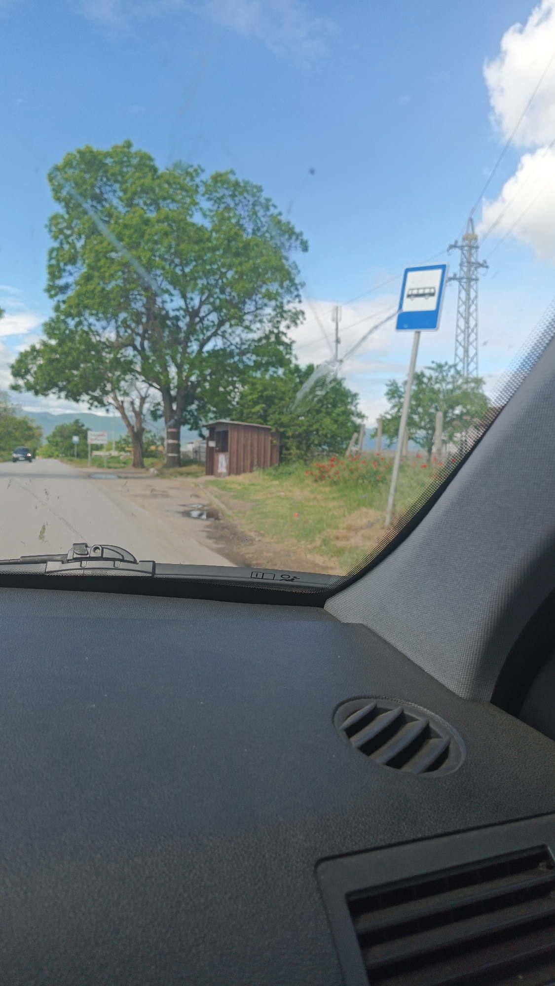 A view from inside a vehicle shows a road scene in Boliartsi, Bulgaria. Visible through the windshield is a two-lane road with a large tree on the left. A small, wooden bus shelter is adjacent to the tree. A bus stop sign is on the right side of the road, near a utility pole. Red flowers are visible in the grass. The vehicle's dashboard is prominent in the foreground, featuring a circular air vent.