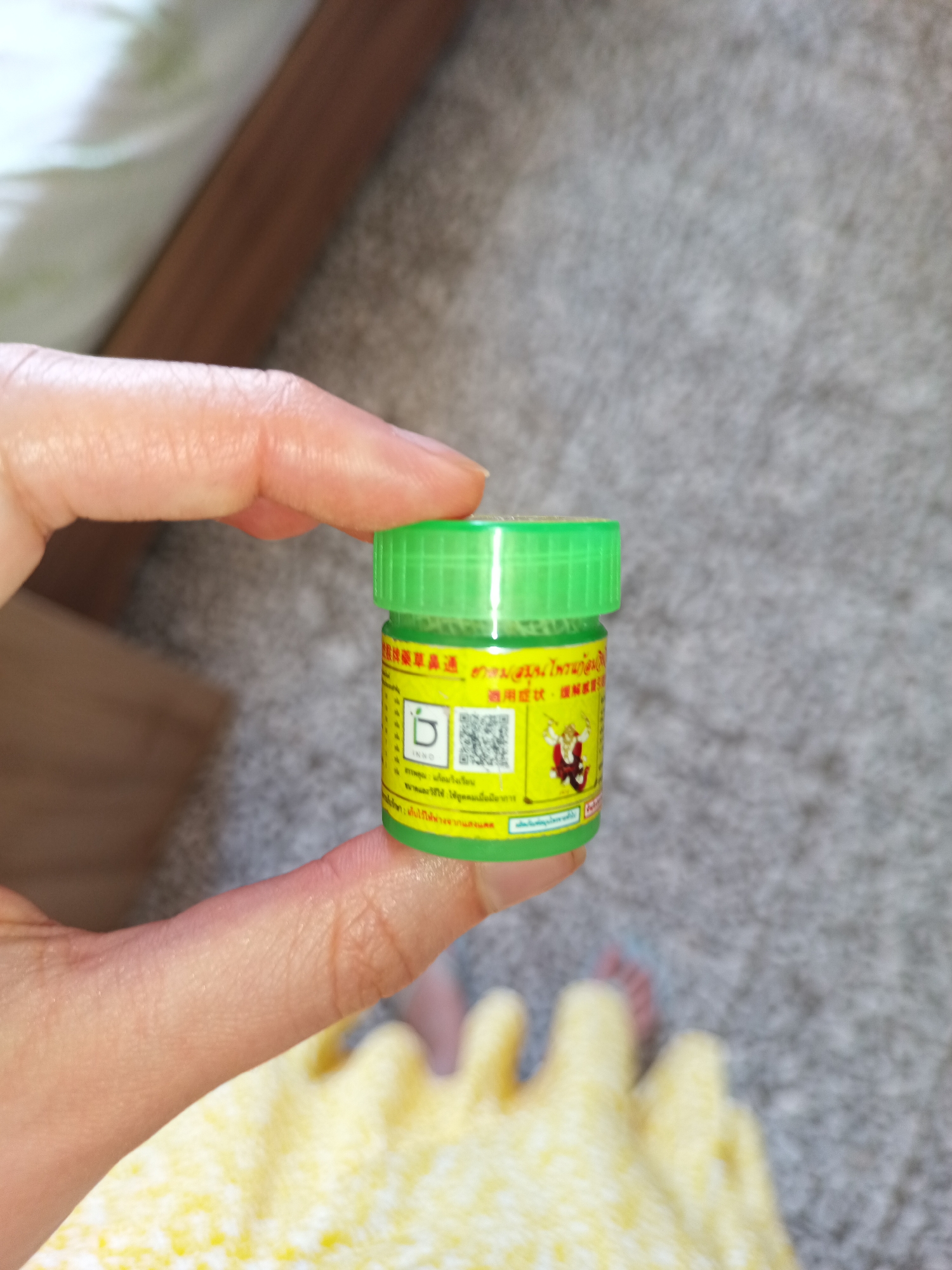 A hand holds a small green jar with yellow labeling in Thai and Japanese. The label also contains an illustration of a monkey or gnome-like character. The background shows a neutral-colored carpet, a wooden frame, and a partially visible person in a yellow dress. The location is not identifiable as Catania, Italy based solely on the visual information provided.