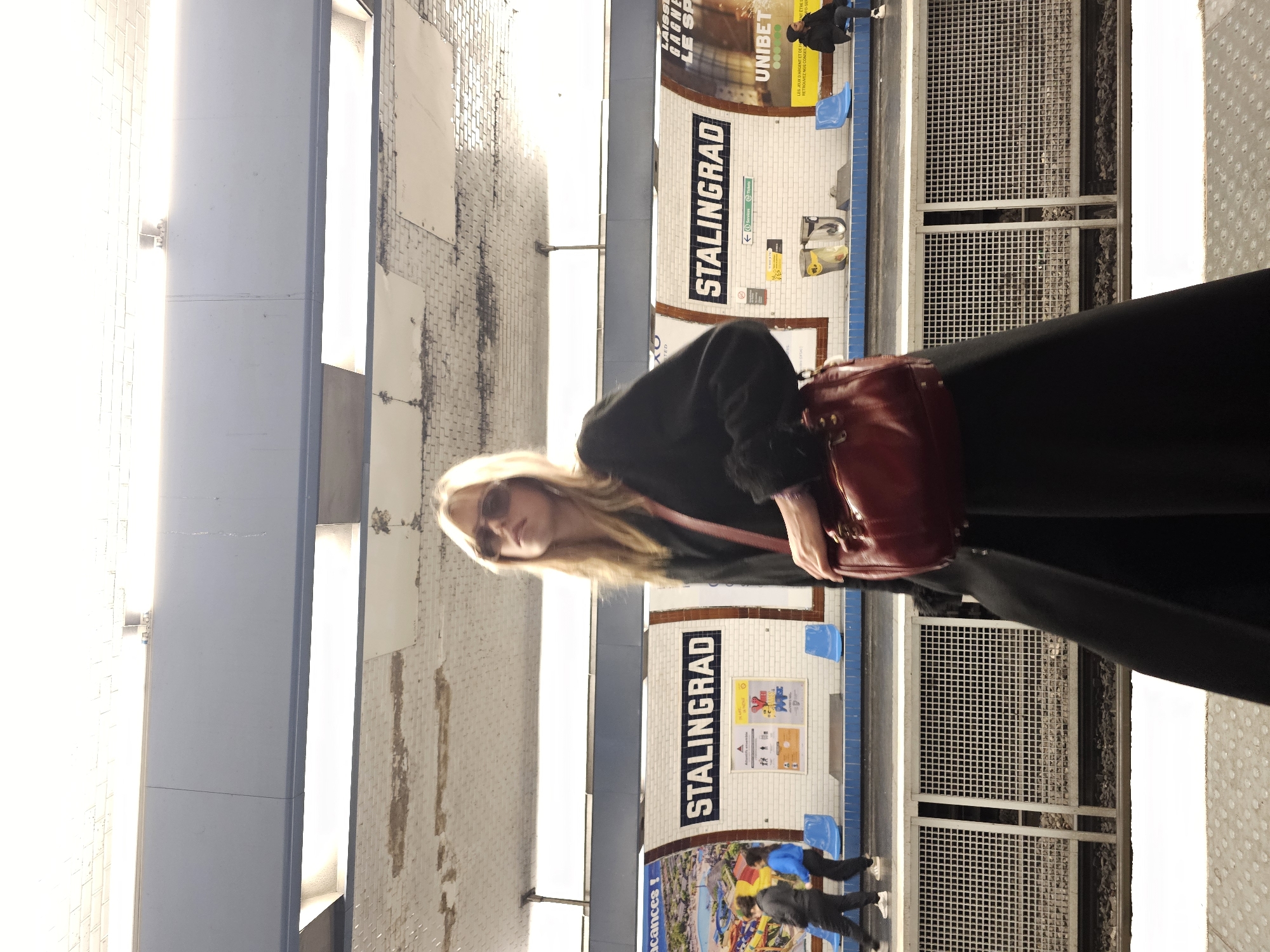 Here is a description of the image:
The image depicts a scene at the Stalingrad metro station in Paris, France. A woman with blonde hair and sunglasses is in the foreground, carrying a red purse. Behind her, the station's tiled walls are visible, marked with "STALINGRAD" signs and advertisements. Blue benches are visible. A person is seated on one of the benches. There are people in the background waiting at the platform edge. The lighting appears to be bright and artificial.