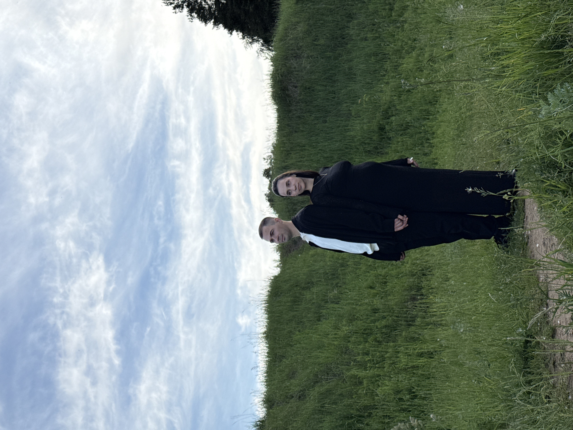 The image displays two individuals, a man and a woman, standing outdoors in a natural landscape, likely in Chaikovsky, Russia. They are positioned in the mid-ground, with a vast green grassy field stretching across the foreground and mid-ground. A narrow dirt path is visible along the right side of the frame, winding through the grass. In the background, a horizon line separates the grassy hill from a expansive sky filled with soft, scattered white clouds. A dark treeline or bush line is faintly visible along the horizon, particularly on the upper left side.
The man, situated on the left, wears a long black garment, possibly a robe or coat, with a prominent white collar or shirt visible underneath. He has short hair and a neutral expression, looking slightly off-camera. The woman stands slightly behind and to the right of the man, also dressed in a long black garment. She has dark hair and a neutral expression, looking directly towards the viewer.
The scene is captured during daytime, under a partly cloudy sky, which provides diffuse and even lighting without strong shadows. The overall setting is rural or semi-rural, characterized by open fields and natural vegetation. The individuals appear to be posing for a photograph. No text is visible within the image.