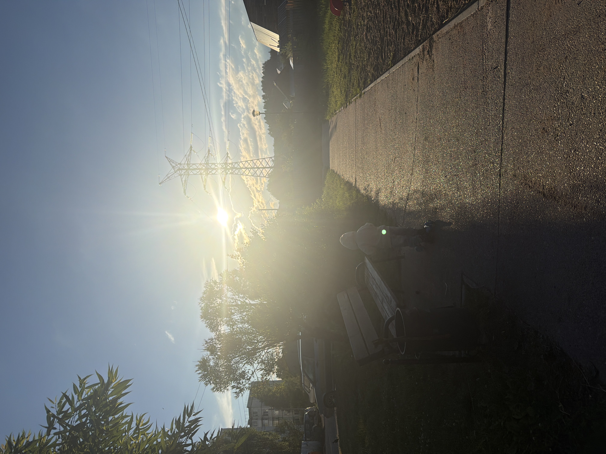 A low-angle, bright sun shines from the left, illuminating a paved walkway in a park. A child wearing a light-colored hooded outfit and riding a scooter is positioned on the walkway, facing away from the camera. To the left of the walkway, a wooden park bench and a trash can are visible, partially obscured by shadows and vegetation. In the background, trees, buildings, power lines, and a large electrical tower are present under a blue sky with scattered clouds. The scene appears to be set in an outdoor urban or suburban environment, possibly a residential area in Chaikovsky, Russia, given the architectural style of the buildings and the presence of the power infrastructure.