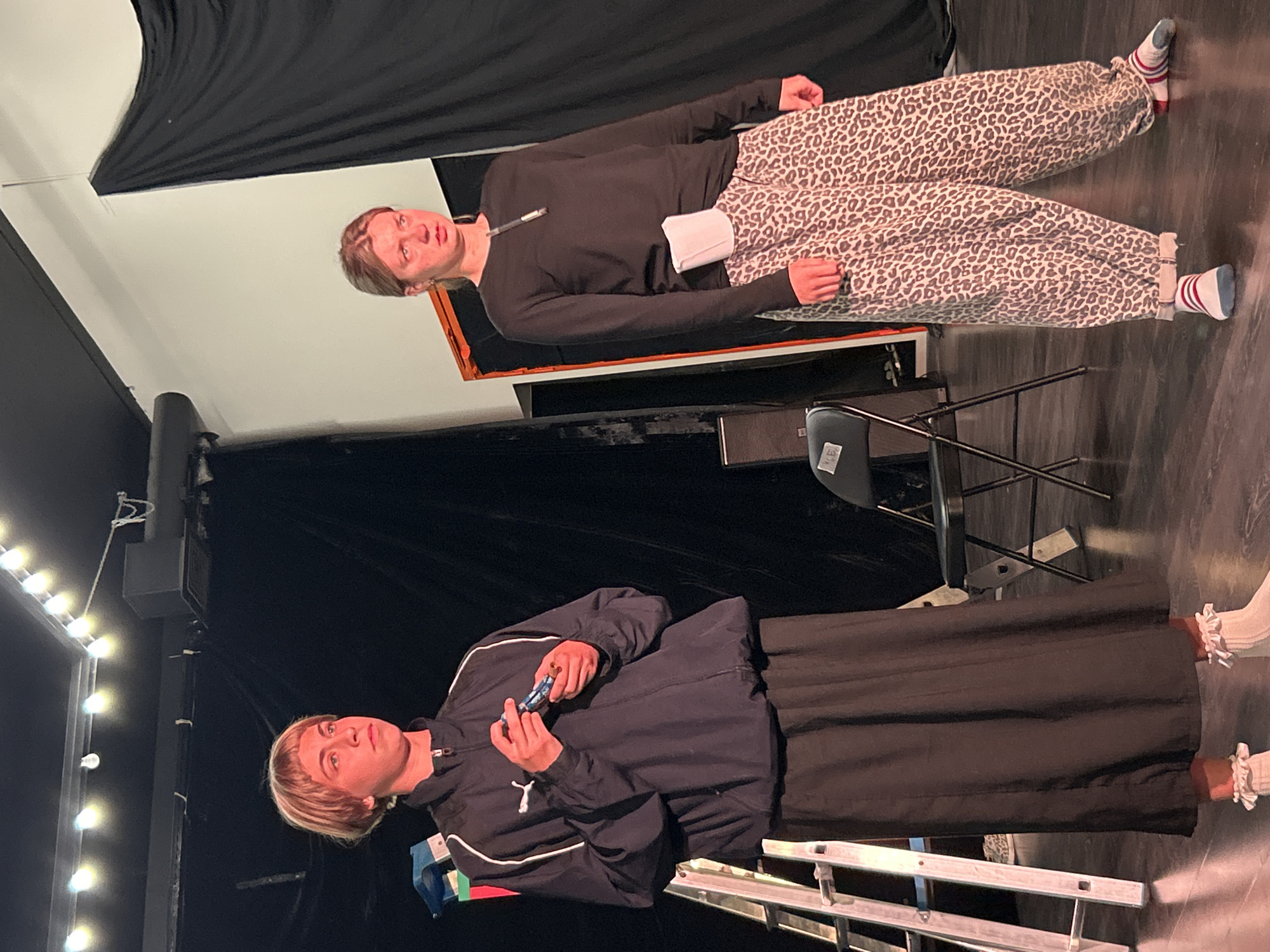Two people stand on a dark stage with a wooden floor. The person on the left, wearing a dark blue jacket over a dark skirt and white frilly socks, holds a small blue object. This person has short blonde hair and is looking to the left. The person on the right wears a black long-sleeved shirt and leopard-print pants, along with striped socks. They are holding a pen with a white clip and looking forward with a distressed expression. Behind them, a black curtain hangs, and to the left, a silver ladder is visible. A chair with a black seat and silver legs is positioned to the right of the ladder. String lights illuminate the upper part of the stage. The scene appears to be a theatrical performance.