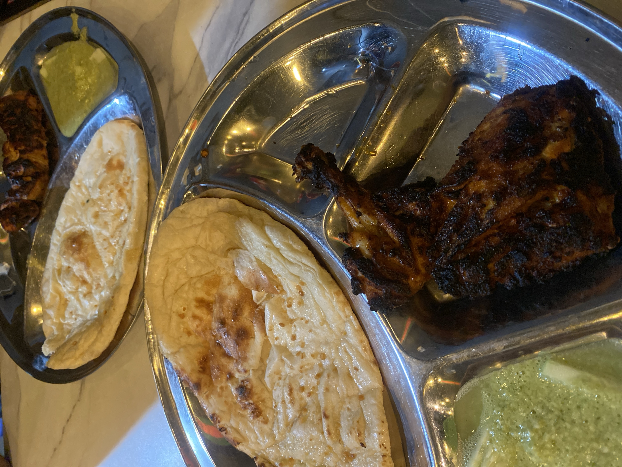 A high-angle shot shows two metal serving dishes, each divided into three sections, containing food, likely in Dhaka, Bangladesh. The left dish contains several pieces of dark meat, a light-colored flatbread, and a green sauce. The right dish also holds a flatbread and green sauce, alongside a single, large piece of dark, grilled meat. The overall environment suggests a restaurant setting.