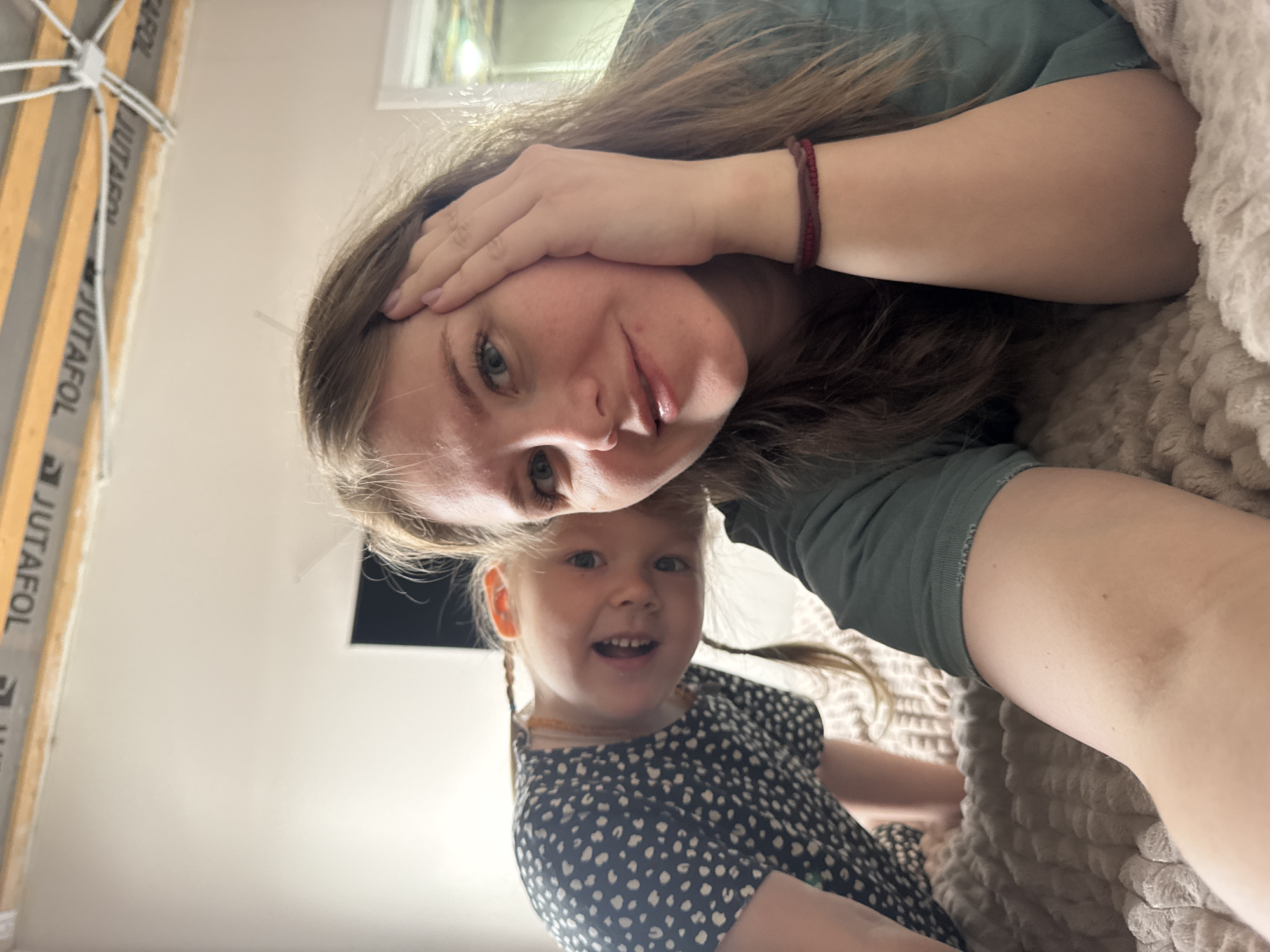 The image depicts a woman and a young girl, likely a mother and daughter, lying down indoors. The woman is fair-skinned with light brown hair and is wearing a green shirt. She is touching her forehead with her hand and looking at the camera. The girl has blonde hair styled in pigtails and is wearing a dark blue dress with small white spots. She is smiling and also looking at the camera. They are lying on a textured, light brown blanket. The background shows a light-colored wall, part of a window, and a piece of construction material (labeled "JUTAFOL"). The specific location in Gornyi, Dvurechenskoe S/P, Russia, cannot be determined from the image.