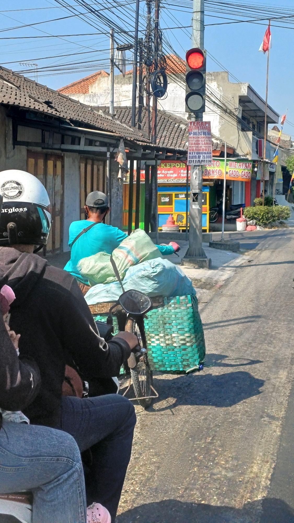 The image depicts a street scene in Delanggu, Indonesia, on a sunny day. In the foreground, two people are seated on a motorcycle, with their backs to the camera. The rider wears a black hoodie and dark pants, while the passenger in front wears light blue jeans. The motorcycle is equipped with a large, green woven basket on the back, loaded with several large, light-colored sacks.
A man wearing a turquoise shirt and a grey baseball cap is riding another motorcycle in front of them. His motorcycle also carries a large amount of cargo in a similar green woven basket, filled with light-colored sacks. He is looking ahead, and his right arm is visible, holding onto the handlebar or a bag.
In the background, a traffic light with a red light illuminated stands prominently. Next to it, a pole with a dense network of electrical wires is visible. Several buildings with tiled roofs line the street, with some shops displaying signs. One sign appears to be for "Mie Ayam Bakso Gado-Gado". A red and white flag is flying on a pole to the right of the traffic light. The road surface is asphalt, with visible cracks and gravel. Shadows are cast on the road, indicating the bright sunlight. The overall environment suggests a typical urban or semi-urban street in Indonesia.