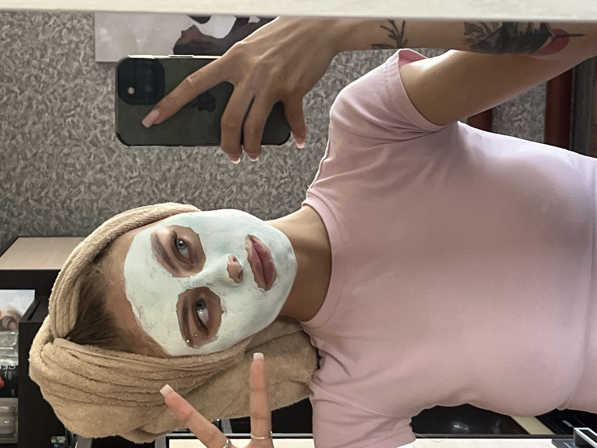 The image shows a young woman with a towel wrapped around her head and a face mask on her face. She is taking a selfie in a mirror, holding a smartphone in her right hand. She is wearing a pink t-shirt. Her left hand is raised, and she is making a peace sign. The background appears to be a room with a textured wall and some shelves with objects on them. The image does not provide any information that would allow for the determination of its location in the city of Фоки, Russia.