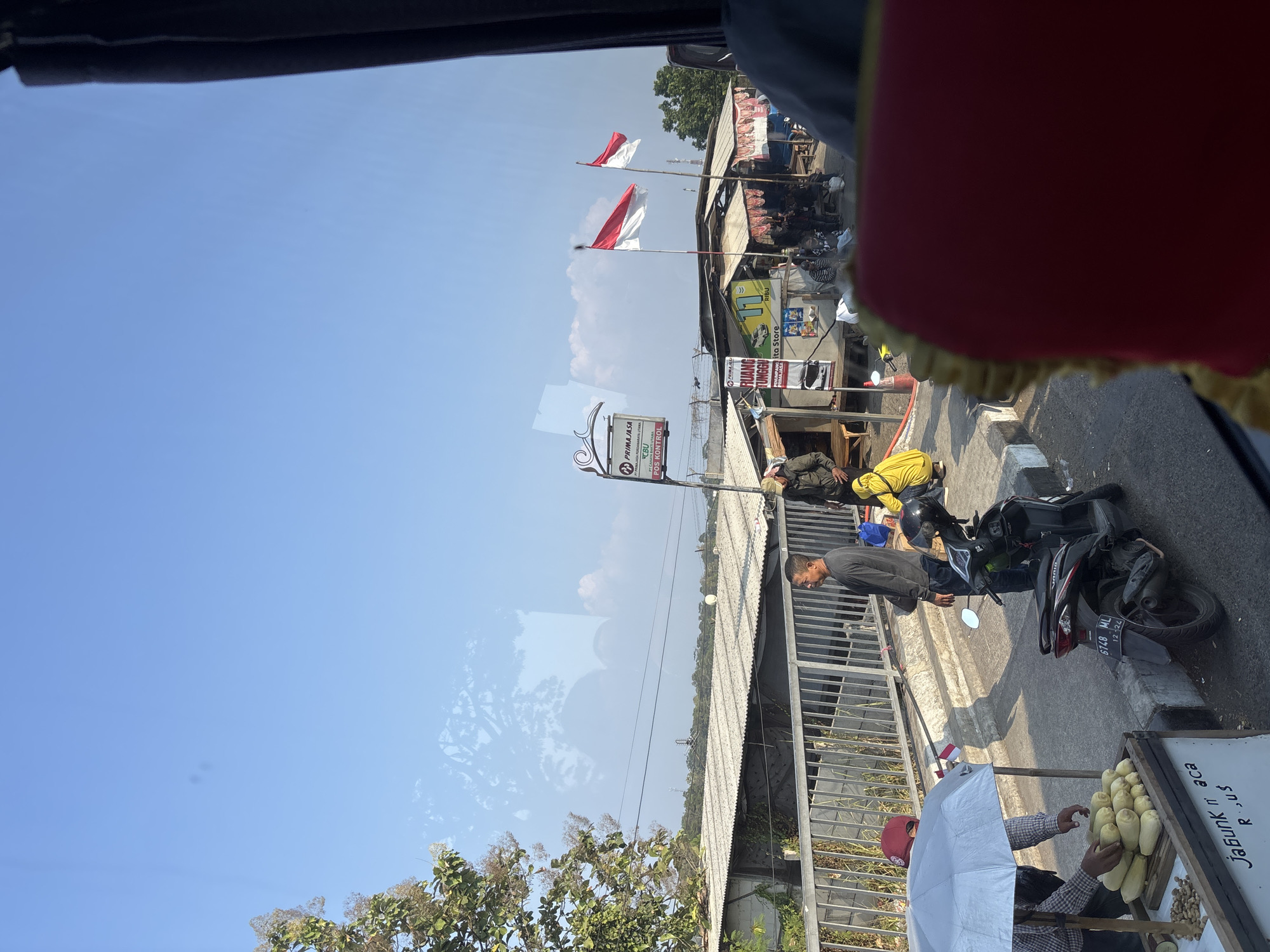 This image depicts a street scene in Cikampek, Indonesia, possibly near a bus station or transportation hub. The foreground shows a vendor under a white umbrella selling corn on the cob. A motorbike is parked beside him, and a person in a red cap is interacting with the vendor. In the middle ground, several buildings with corrugated metal roofs are visible, along with a metal railing and a pedestrian walkway. Two Indonesian flags are flying from poles in front of a roadside stall. A group of people are gathered near the stalls, some sitting and others standing. The background shows a clear blue sky with scattered clouds and some vegetation. The overall impression is one of a busy, functional urban environment.