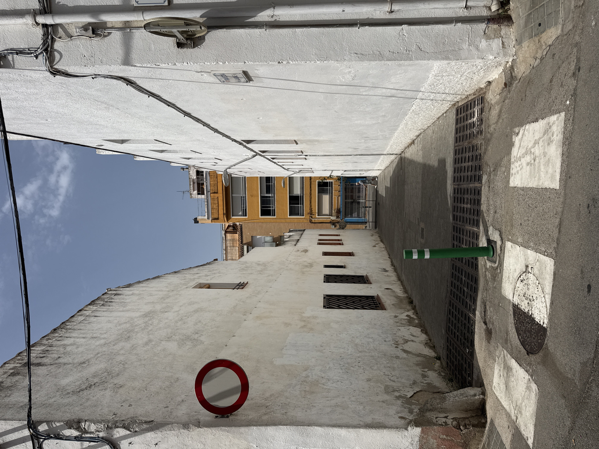 The image depicts a narrow alleyway in the city of Blanes, Spain. The alley is flanked by tall, aged buildings with white, textured facades. The building on the left shows signs of wear, with peeling paint and patches of exposed concrete, and features a circular red and white traffic sign. The building on the right is also white and appears more recently maintained, with visible pipes and electrical conduits running along its exterior.
The ground of the alley is paved with asphalt and features a pedestrian crossing marked with white paint. A metal grate covers a drainage channel in the center of the alley, and a green traffic bollard with white stripes stands to the right of the grate.
In the background, the alley opens up to reveal more buildings, including a brightly colored yellow structure with multiple windows and balconies. A person is visible walking away from the camera towards the end of the alley. The sky is clear blue with a few scattered clouds, indicating a sunny day. The overall scene is a typical urban setting, characterized by its architecture, infrastructure, and the presence of pedestrian and vehicular pathways.