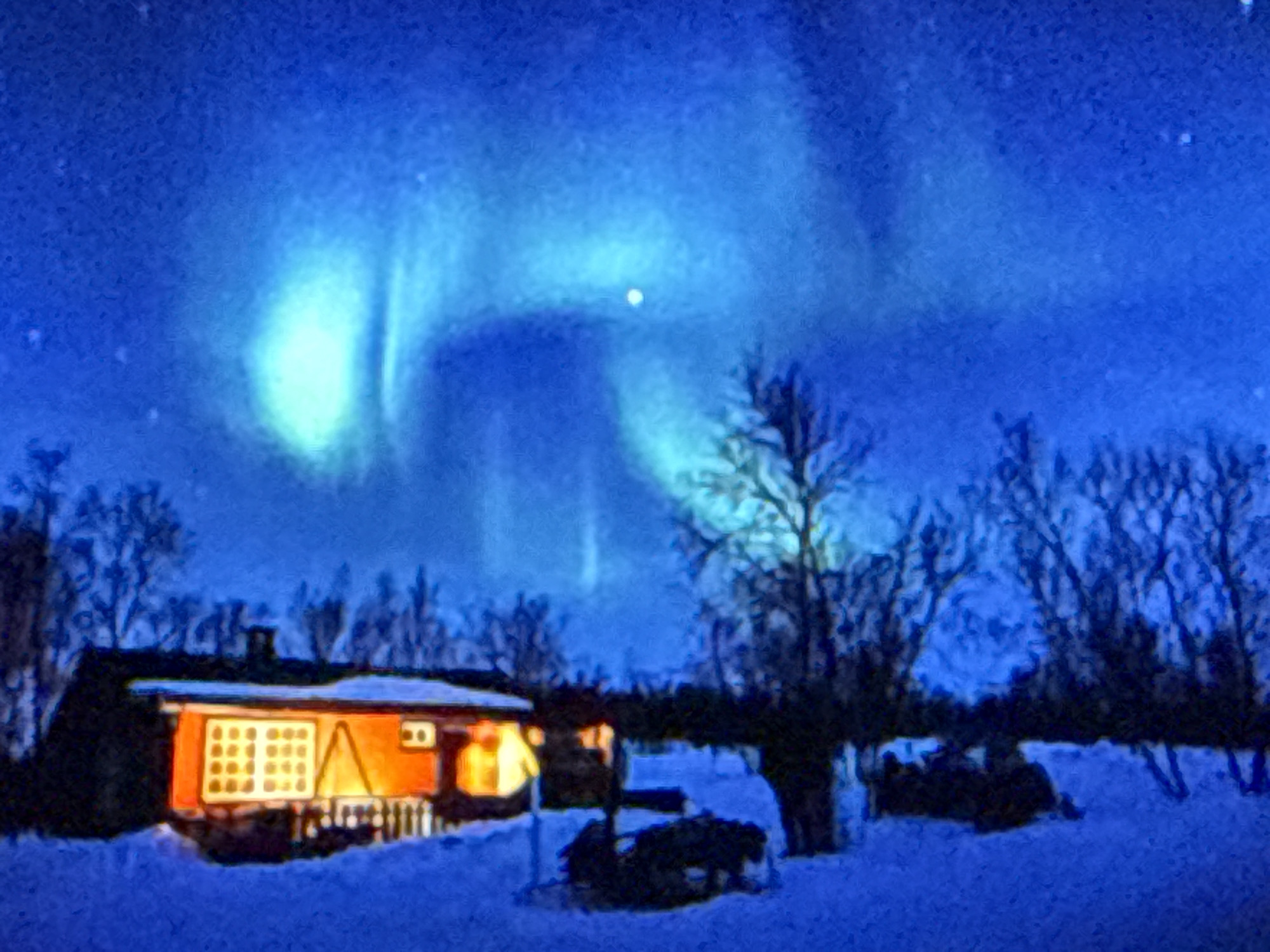 The image depicts a night scene in a snowy landscape, possibly in a rural area, under a vibrant aurora borealis. A small, red-orange house with glowing yellow windows is visible on the left side of the frame, with snow covering the ground and the roof of the house. Bare trees with dark branches are scattered throughout the scene, some in front of and behind the house, contributing to a stark, wintery atmosphere. The sky is dominated by the ethereal green and blue hues of the aurora, with streaks and waves of light illuminating the darkness. A small, bright star or planet is also visible in the sky, to the right of the brightest part of the aurora. The ground is blanketed in deep blue snow, and some dark, indistinct shapes, possibly trees or snowdrifts, are present in the foreground. The overall impression is one of a remote and tranquil winter night, with the natural spectacle of the northern lights providing a dramatic backdrop to the human dwelling.
