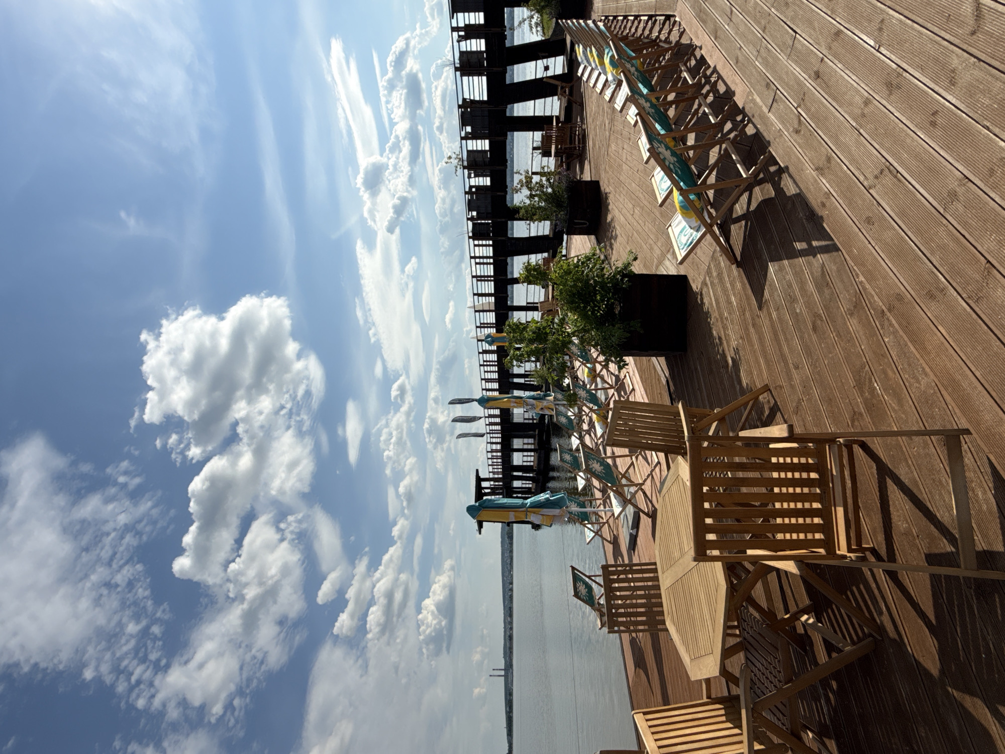 A waterfront dining and lounging area is depicted on a wooden deck overlooking a body of water. The deck is furnished with several wooden tables and chairs, as well as rows of deck chairs with striped cushions. Two large umbrellas, one yellow and blue striped, and another yellow, blue, and white striped, are set up near the deck chairs. Planters with green foliage are strategically placed on the deck. In the background, a dark wooden pier extends over the water. Beyond the pier, a vast expanse of water stretches towards a distant horizon, where a city skyline with industrial chimneys is visible under a partly cloudy blue sky. The scene suggests a recreational area, possibly a restaurant or cafe, situated in the city of Votkinsk, Russia.
