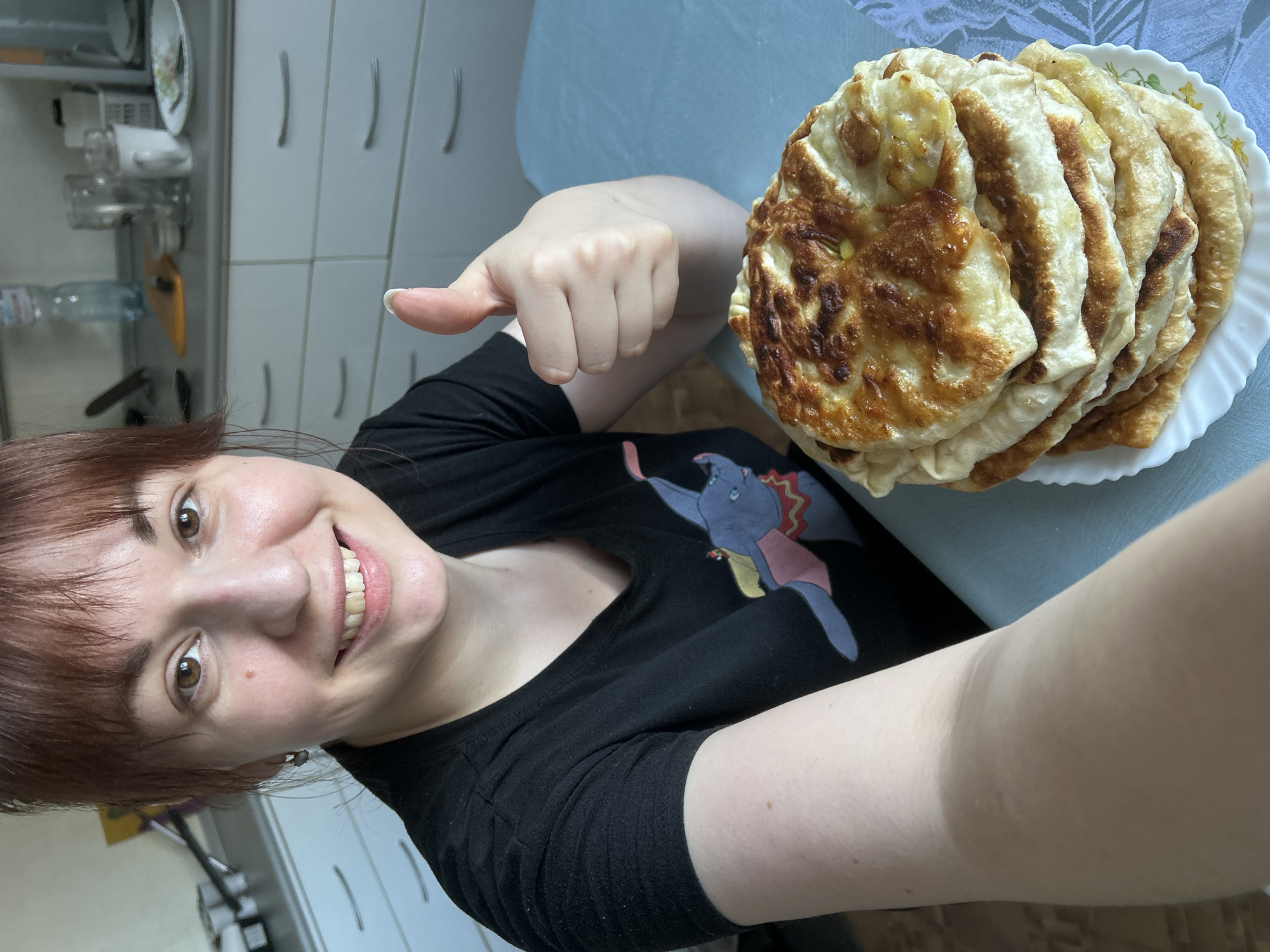 The image depicts a female in a domestic kitchen environment, identified as being in Фоки, Russia. The female, with reddish-brown hair and bangs, is positioned in the foreground, smiling directly at the camera. She wears a black short-sleeved t-shirt featuring a colorful graphic of an elephant. Her left hand is raised, displaying a "thumbs-up" gesture. To her right, a stack of six golden-brown, pan-fried flatbreads, possibly filled, rests on a white plate with a subtle floral pattern, placed on a light blue tablecloth. The background reveals a kitchen area with light-colored cabinets below a countertop. Visible on the countertop are a clear plastic bottle of water, a clear drinking glass, a white mug, a white plastic basket, and an orange cutting board, along with other indistinct kitchen items.