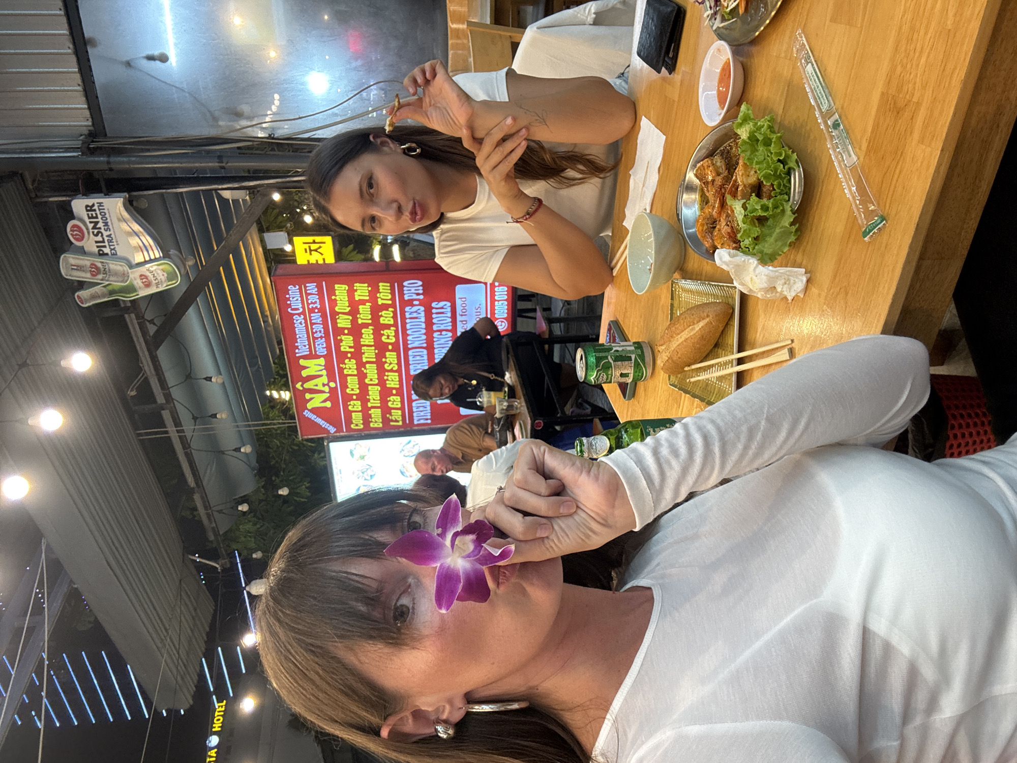 This image captures a nighttime scene inside an open-air Vietnamese restaurant in Đà Nẵng, Vietnam.
In the foreground, two women are seated at a light-colored wooden table. The woman on the left has medium brown hair with bangs and wears a white long-sleeved top. She holds a purple orchid flower to her nose/mouth area, partially obscuring her face, and looks directly at the camera. She wears a silver hoop earring and a smaller silver ball stud earring on her left ear. To her right, a green Heineken beer bottle is visible.
The woman on the right has long brown hair and wears a white short-sleeved t-shirt. She holds a small piece of food to her mouth, with her lips slightly puckered, and looks towards the camera. She wears a gold hoop earring on her right ear, a thin red bracelet on her left wrist, and a visible tattoo on her right forearm.
On the table between them, a green Heineken beer can, a light blue ceramic bowl, a black wallet, white paper napkins, and a small white bowl containing an orange dipping sauce are arranged. A silver metal platter holds several cooked prawns/shrimp served on a bed of green lettuce. A clear plastic-wrapped package containing chopsticks and a toothpick lies on the table. To the left, a baguette rests on a white rectangular plate alongside a pair of wooden chopsticks.
In the background, overhead bare light bulbs illuminate the scene. A large red sign with white text advertises "Vietnamese Cuisine," listing dishes such as "Cơm Gà," "Phở," and "Mỳ Quảng," with operating hours "OPEN: 9:30 AM - 3:30 AM." Above this, a "PILSENER EXTRA SMOOTH" beer sign is mounted, alongside a white sign featuring Korean text and the word "Restaurants." Other patrons are seated at adjacent tables; a man in a brown shirt is visible to the left background, and a woman in a black top is focused on a device at her table further back. Beyond the restaurant's covered area, a distant building with illuminated blue horizontal strip lighting is visible against the dark night sky, partially revealing "STA HOTEL" text. The ceiling structure consists of corrugated metal roofing with exposed wiring.