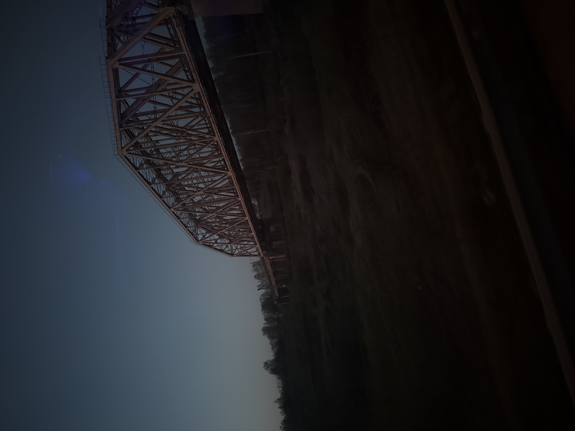 The image shows a steel truss bridge spanning a river in Yakhroma, Russia, likely a railway bridge. The camera is positioned looking upward at the bridge. The sky is a gradient of blue to twilight. Trees line the river bank. The image is rotated and somewhat underexposed.
