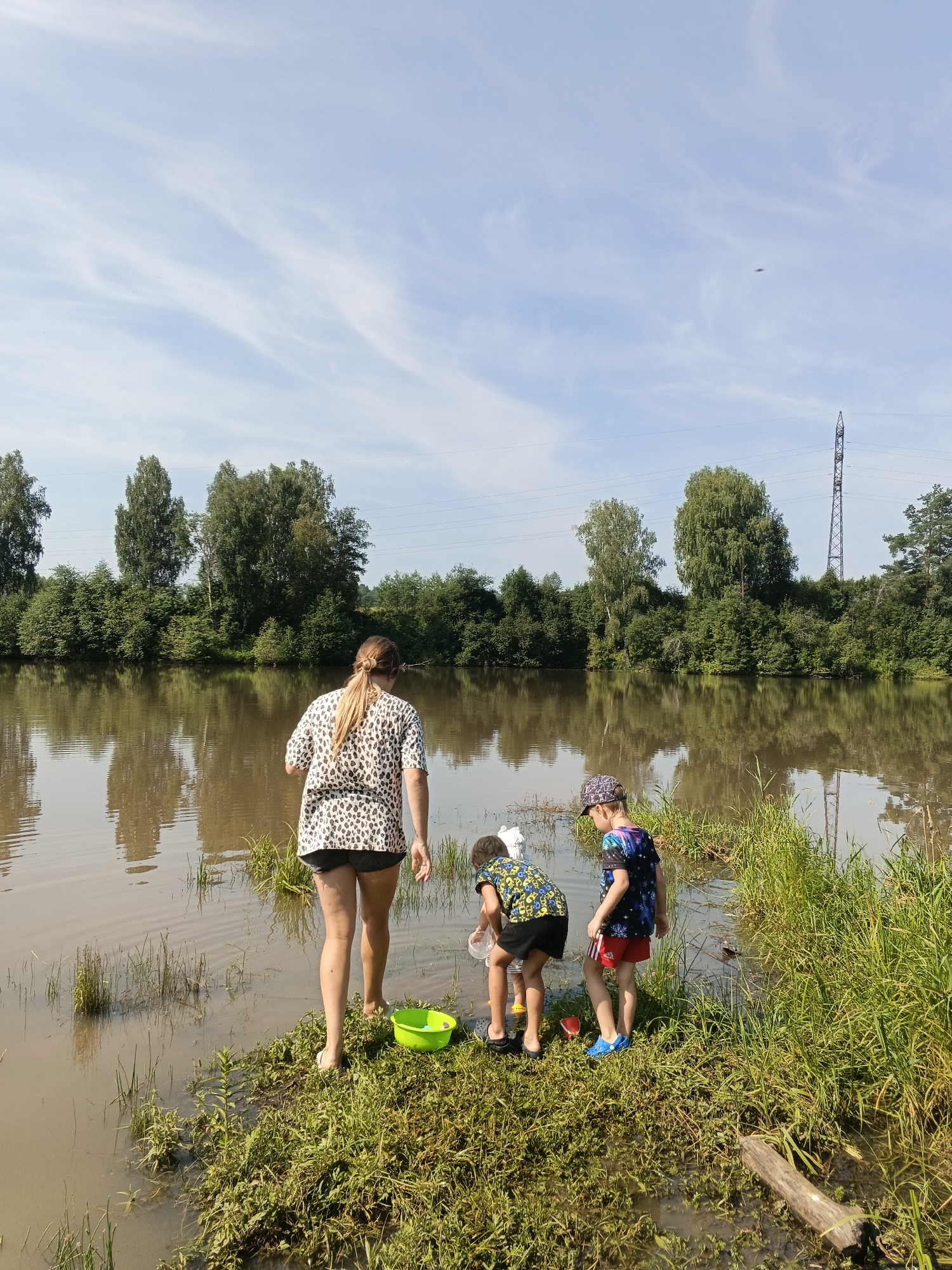 The image shows a woman and two children by a body of water. The woman, with her back to the camera, is wearing a black and white patterned top and dark shorts. She is standing in shallow water. To her right, two children are also by the water. One child, wearing a dark t-shirt with a colorful pattern and red shorts, is facing away from the camera and looking down at the water. The other child, wearing a dark patterned t-shirt and black shorts, is also facing away from the camera and appears to be reaching into the water. There is a green bucket near the woman and a clear container in the water near the children. The background consists of trees and a bright blue sky with some wispy clouds. In the distance, a power line tower is visible. The water is calm and reflects the trees and sky. The ground near the water is covered in green grass and some debris.