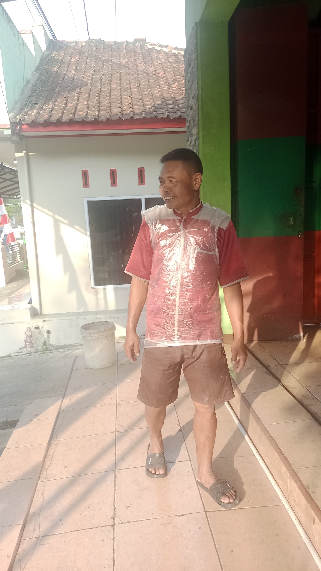 A man, appearing to be of Indonesian descent, stands on a tiled walkway in front of a building. He has short, dark hair and is smiling. He is wearing a red short-sleeved shirt with a clear plastic vest over it, and brown shorts. He is wearing dark-colored slides on his feet. The building behind him has a tiled roof and a light-colored exterior with red accents. To the right of the man, a portion of the building is painted in vertical stripes of green and dark red. The ground in front of the building is tiled, and a light-colored cylindrical object, possibly a bucket, is visible to the left of the man. The scene is illuminated by sunlight, casting shadows on the ground. The location is identified as Karyamandala, Indonesia.