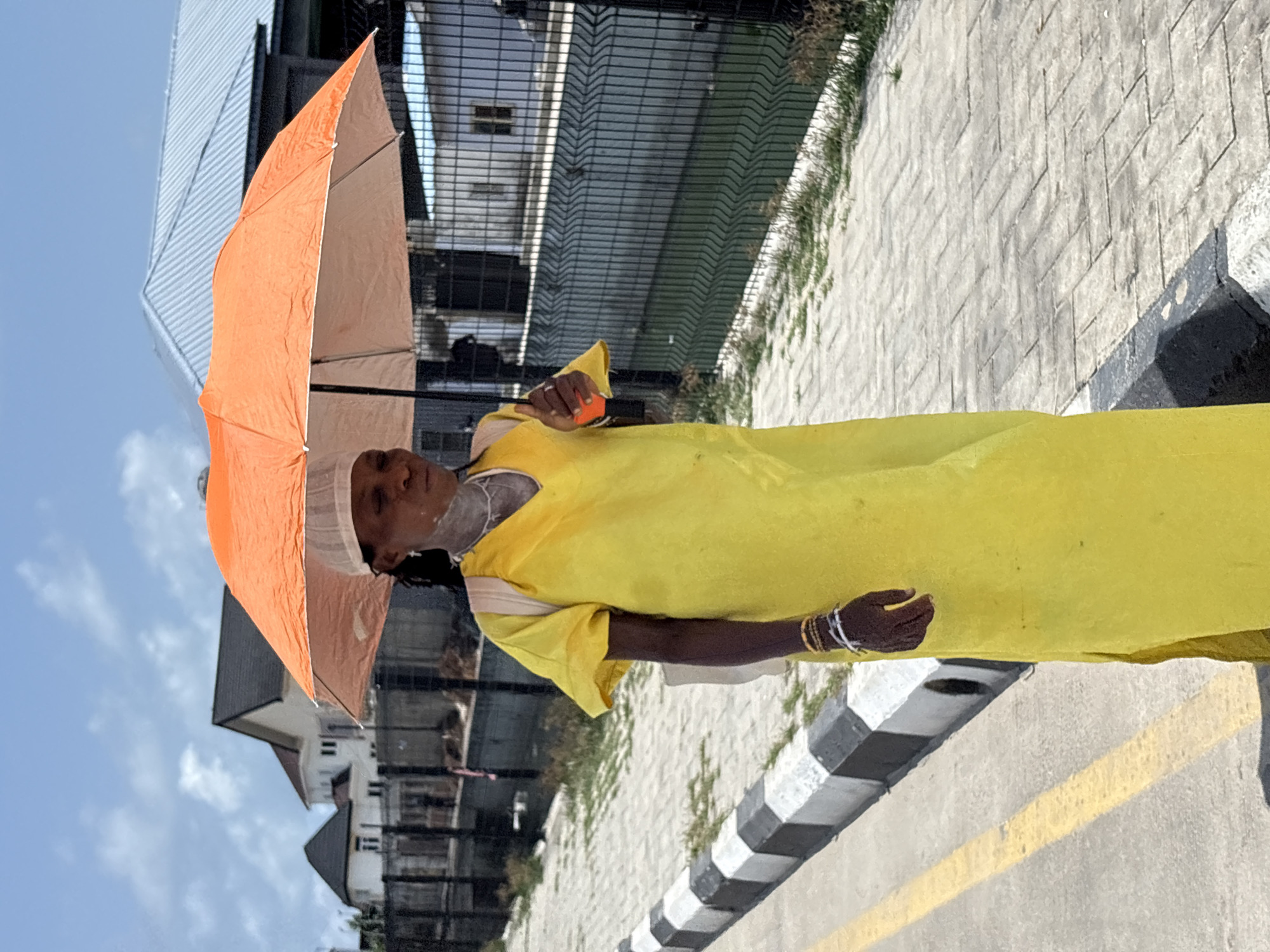 The image, taken in Igboefon, Nigeria, captures an individual, likely a woman, standing outdoors on a bright day. She is positioned towards the center of the frame, facing slightly left and looking upwards. She wears a long, flowing, bright yellow dress or tunic, and a light-colored, possibly white, head covering. A distinct white or patterned substance is visible on her neck and upper chest area.
She holds an open, vibrant orange umbrella in her left hand, utilizing it for shade from the intense sunlight. Her right arm is visible, adorned with multiple bangles in gold, white, and black.
The setting features a paved surface in the foreground, leading to a prominent black and white striped curb or barrier that runs horizontally. Beyond this, the ground is composed of light-colored paving blocks or concrete, with a yellow line marking a road surface further in the background. Behind the individual, a dark metal fence is visible, and beyond it, several buildings with light facades and dark roofs can be seen under a clear blue sky with scattered faint clouds. The bright lighting suggests it is daytime, likely midday or early afternoon. No discernible text is visible in the image.