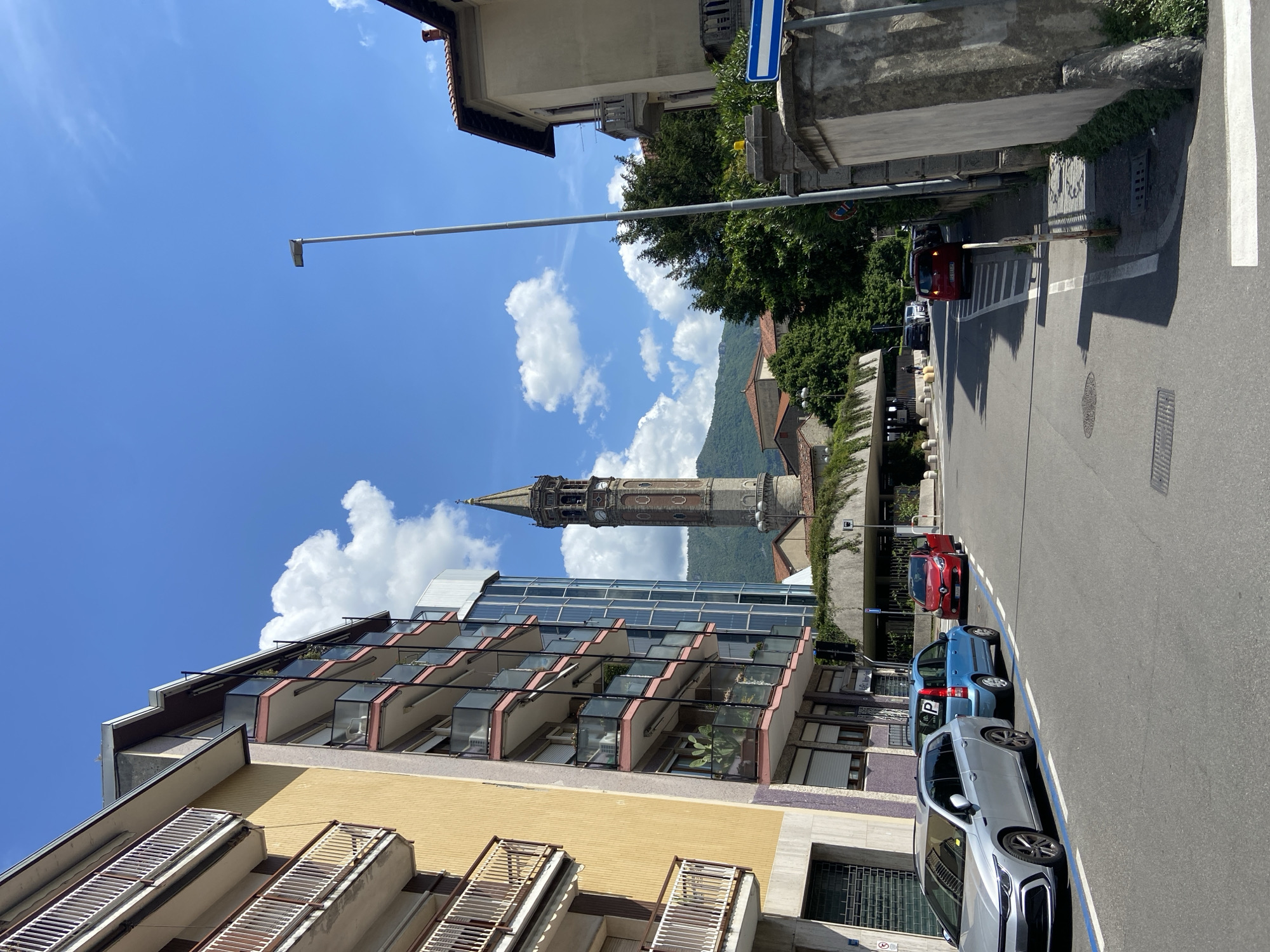The image depicts a street scene in Lecco, Italy, under a bright blue sky with scattered clouds. A tall, ornate bell tower dominates the background, partly obscured by a modern building with glass balconies and a yellow facade. Several cars are parked along the right side of the street, including a gray, blue, and red vehicle. A street sign is visible. The road is marked with a crosswalk. In the background, a tree-covered hillside rises behind the buildings.