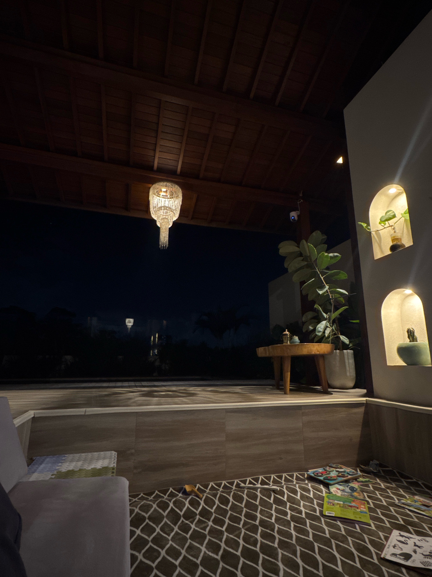 An outdoor covered patio area is depicted at night. The ceiling is constructed of dark wooden beams. A large, ornate glass chandelier hangs from the ceiling. In the background, the dark sky is visible, with some distant lights and faint silhouettes of trees and structures.
On the patio, a grey sofa is positioned to the left. In front of the sofa, on the floor, is a patterned rug with a white diamond design. Scattered on the rug are several children's books and puzzles. To the right of the sofa, a raised platform is visible with a wooden table on it. A potted plant with large green leaves stands next to the table, and behind it, a wall features two arched recessed shelves, each containing a small plant. A security camera is mounted on the ceiling structure.
The scene is located in Mengwi, Indonesia.