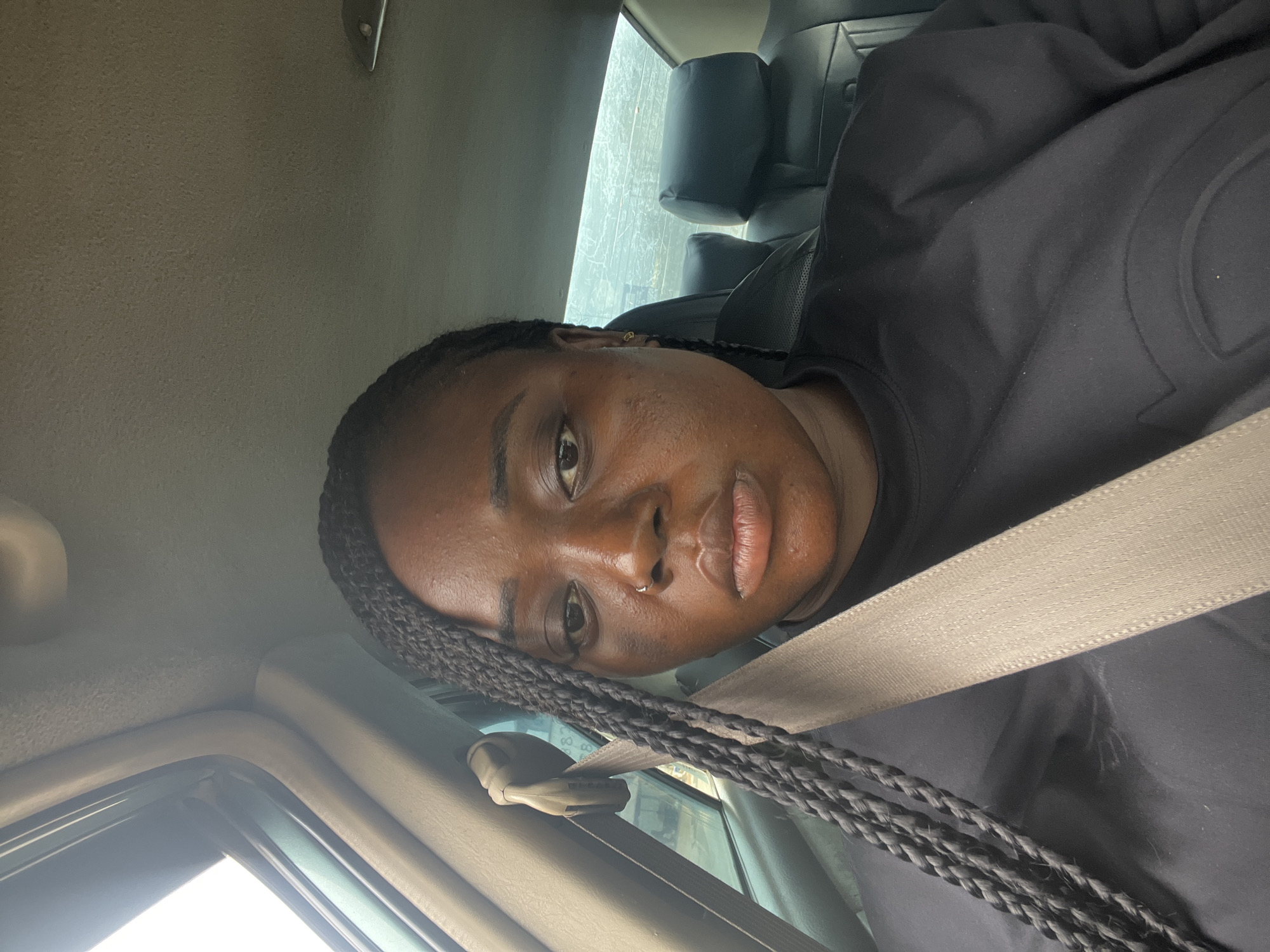 A person is inside a vehicle, wearing a seatbelt, and facing the camera. The person has dark skin and braids and is wearing a black top. The location appears to be Abuja, Nigeria. The interior of the vehicle is visible, including seats and a window.