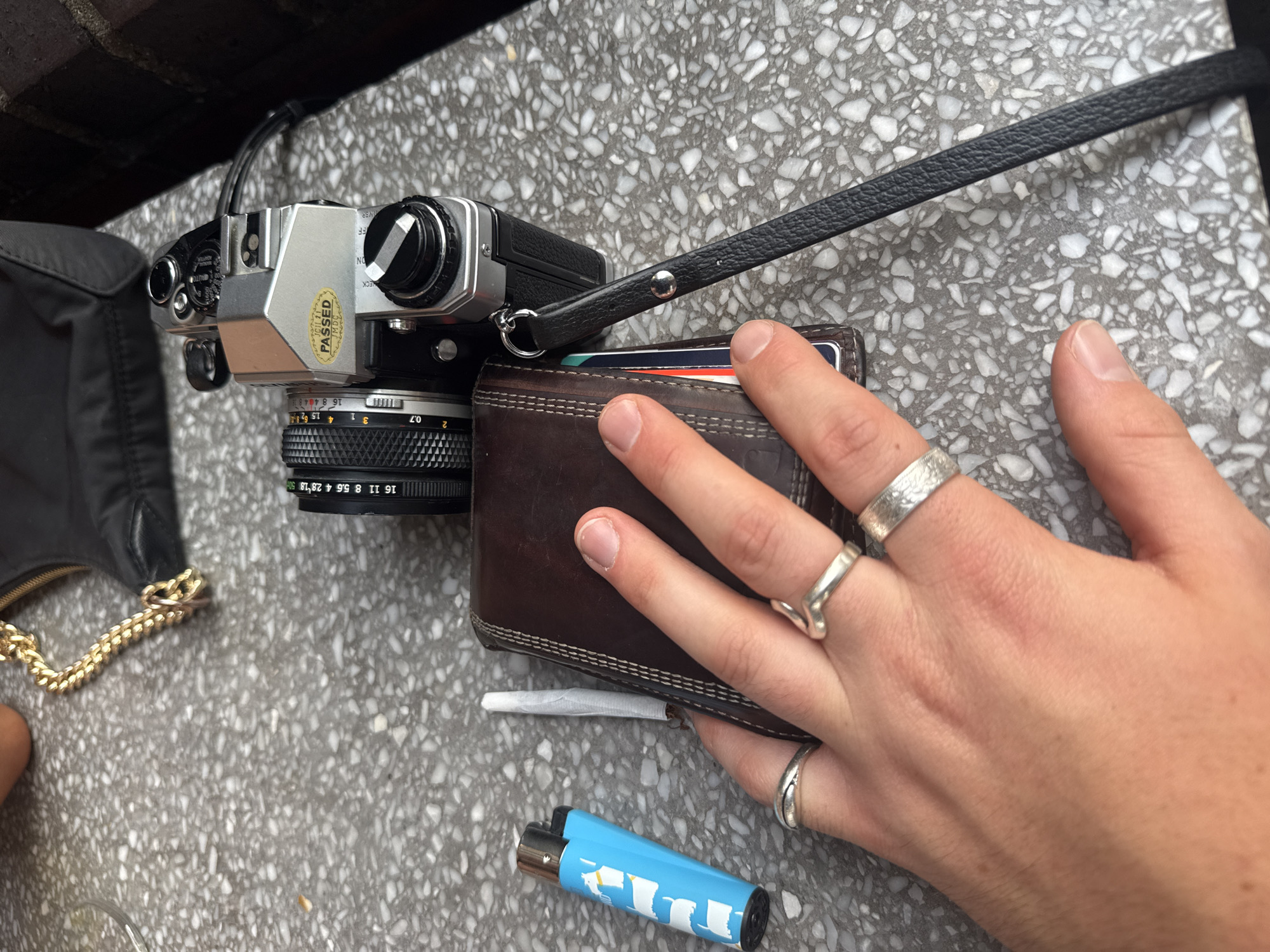 Here's a detailed description of the image:
**Scene:** An outdoor still life, likely on a stone or concrete surface with a speckled, terrazzo-like pattern.
**Objects:**
* **Camera:** A vintage silver and black film camera with a black lens attached. A black strap is connected to the camera.
* **Wallet:** A worn brown leather wallet with cards visible inside.
* **Hand:** A hand rests on the wallet. The hand has multiple silver rings.
* **Joint:** A rolled cigarette or joint is visible near the bottom edge of the wallet.
* **Lighter:** A light blue lighter with a silver top.
* **Bag:** Part of a black bag with a gold chain is visible on the left side.
**People:** No people are fully visible, but a hand is present.
**Actions:** The hand is positioned over the wallet.
**Interactions:** The hand interacts with the wallet. The camera rests in part on the wallet.
**Environment:** The setting appears to be outdoors, possibly on a bench or table. The lighting suggests it's daytime. The location in the city of Unknown, Unknown, cannot be determined.