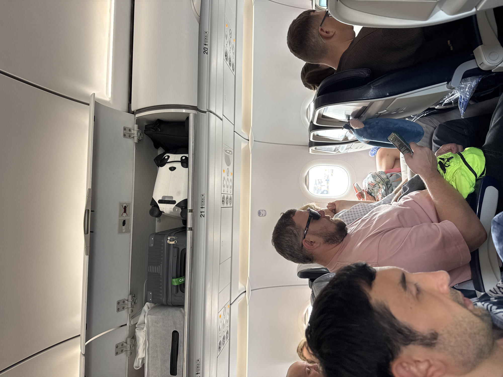 The image depicts the interior of a commercial passenger aircraft cabin, specifically focusing on the overhead bins and several seated passengers. The location is specified as ตำบล ไม้ขาว, Thailand.
In the upper left, an overhead compartment, numbered "21 CBA," is open, revealing multiple items of carry-on luggage. From left to right inside this bin: a light grey fabric soft-sided case, a dark grey hardshell suitcase with vertical ridging and a green tag that partially reads "CAE," and a white object with black accents, possibly a specialized case or another piece of luggage. A black bag is partially visible further right within the adjacent bin section. A safety decal inside the open bin displays symbols for a charging cable and a prohibited crossed-out charging cable with a battery icon. The adjacent overhead bin section to the right, labeled "20 CBA," is closed. Below the closed bin, a "Wi-Fi" symbol is affixed to the cabin wall.
In the mid-ground, a male passenger with a beard, short grey hair, and dark sunglasses is seated, wearing a pink short-sleeved t-shirt. He holds a dark-colored smartphone in his left hand and a bright lime-green fabric item, possibly a small bag or garment, in his right hand, resting on his lap. He has a ring on his right ring finger. His arm rests on the shared armrest.
To the right of the man in the pink shirt, another male passenger with short dark hair and glasses is visible from behind and in profile, wearing a dark brown long-sleeved shirt. His head is turned slightly away from the camera. A blue plush object, resembling a small travel pillow or stuffed animal, hangs from the headrest of the seat in front of him. A clear plastic bag containing an unknown item is partially visible on the floor or seat base in front of this passenger.
In the immediate foreground on the left, the left profile of a male passenger with dark hair and stubble is visible. He is wearing a patterned shirt and looking downwards. Partially visible airplane windows in the background show an indistinct external view, possibly a tarmac or sky. Overhead passenger service units with lights and vents are visible below the closed overhead bins. The aircraft seats are predominantly dark blue with light grey or white frames.