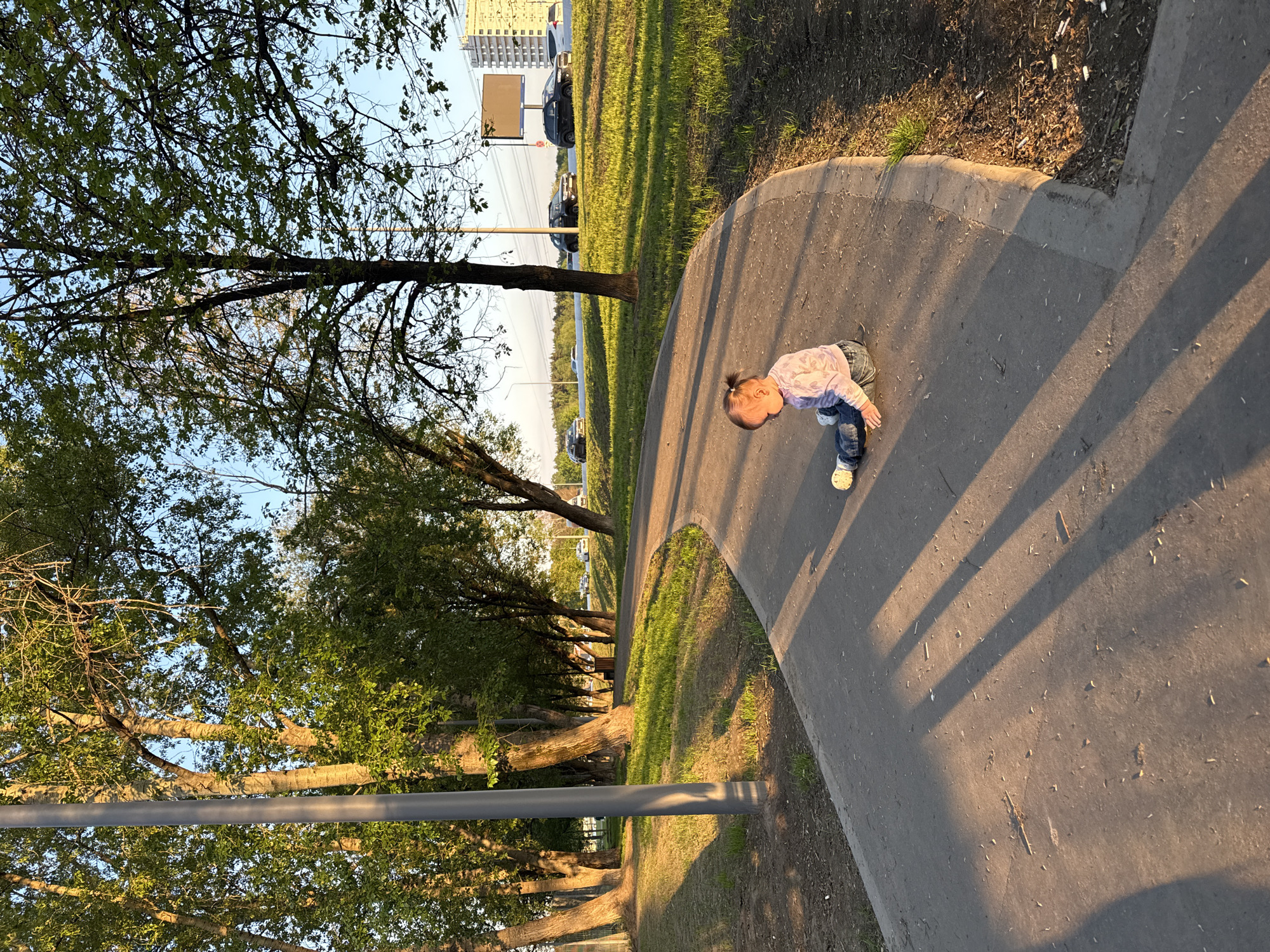 A young child is on a concrete pathway in a park, presumably in Moscow, Russia. The child is wearing a light-colored patterned shirt, blue jeans, and white shoes. The sun casts long shadows on the pathway. The pathway curves and is bordered by a grassy area with trees. A row of trees lines the pathway, and beyond them, there is a road with cars and a building in the distance. A metal pole is visible near the edge of the pathway.
