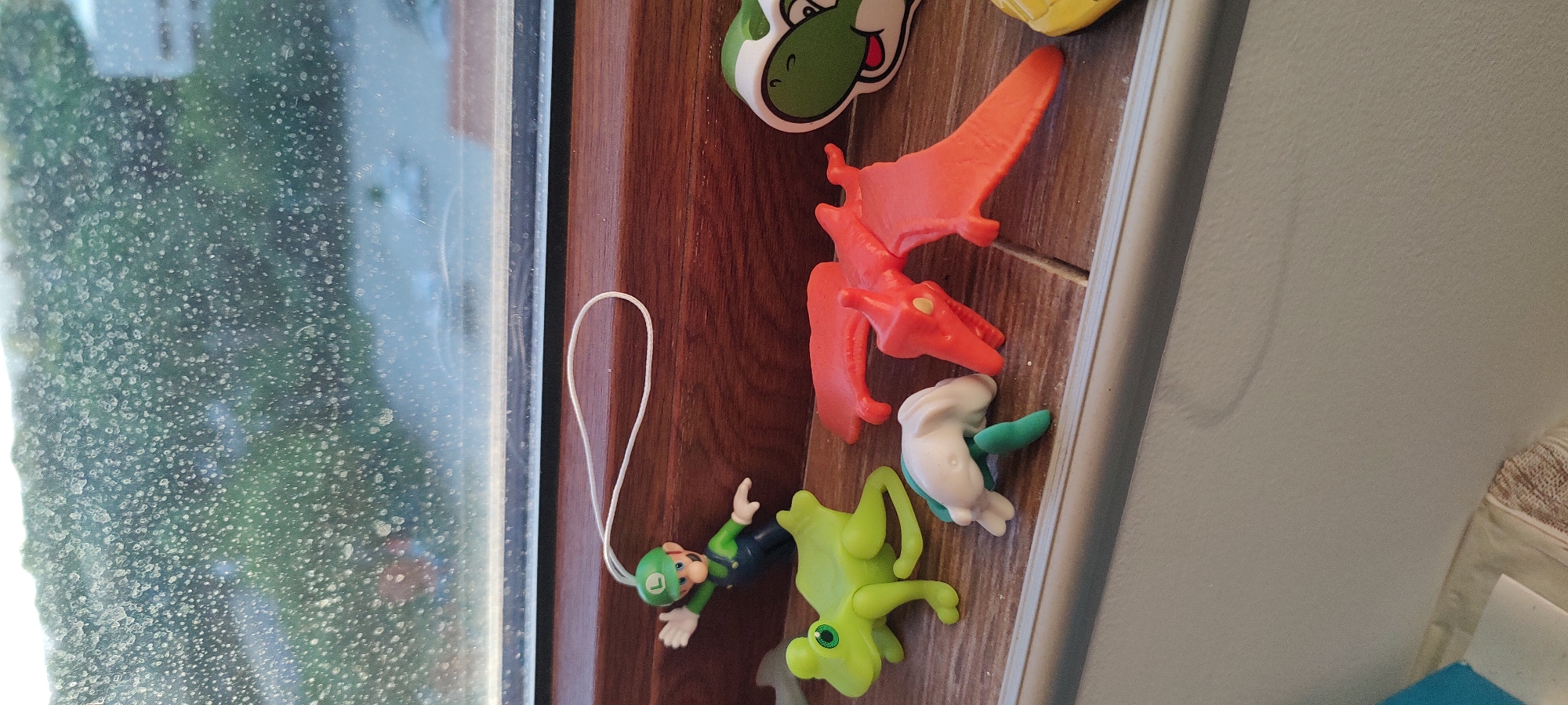 The image shows a collection of toys and a window. The toys consist of a Luigi figurine with a hanging loop, a green frog toy, a white and green animal figure, and an orange pterodactyl. The window is visible on the left, with a view obscured by condensation. A brown wooden surface is present behind the toys. The environment suggests an indoor setting.