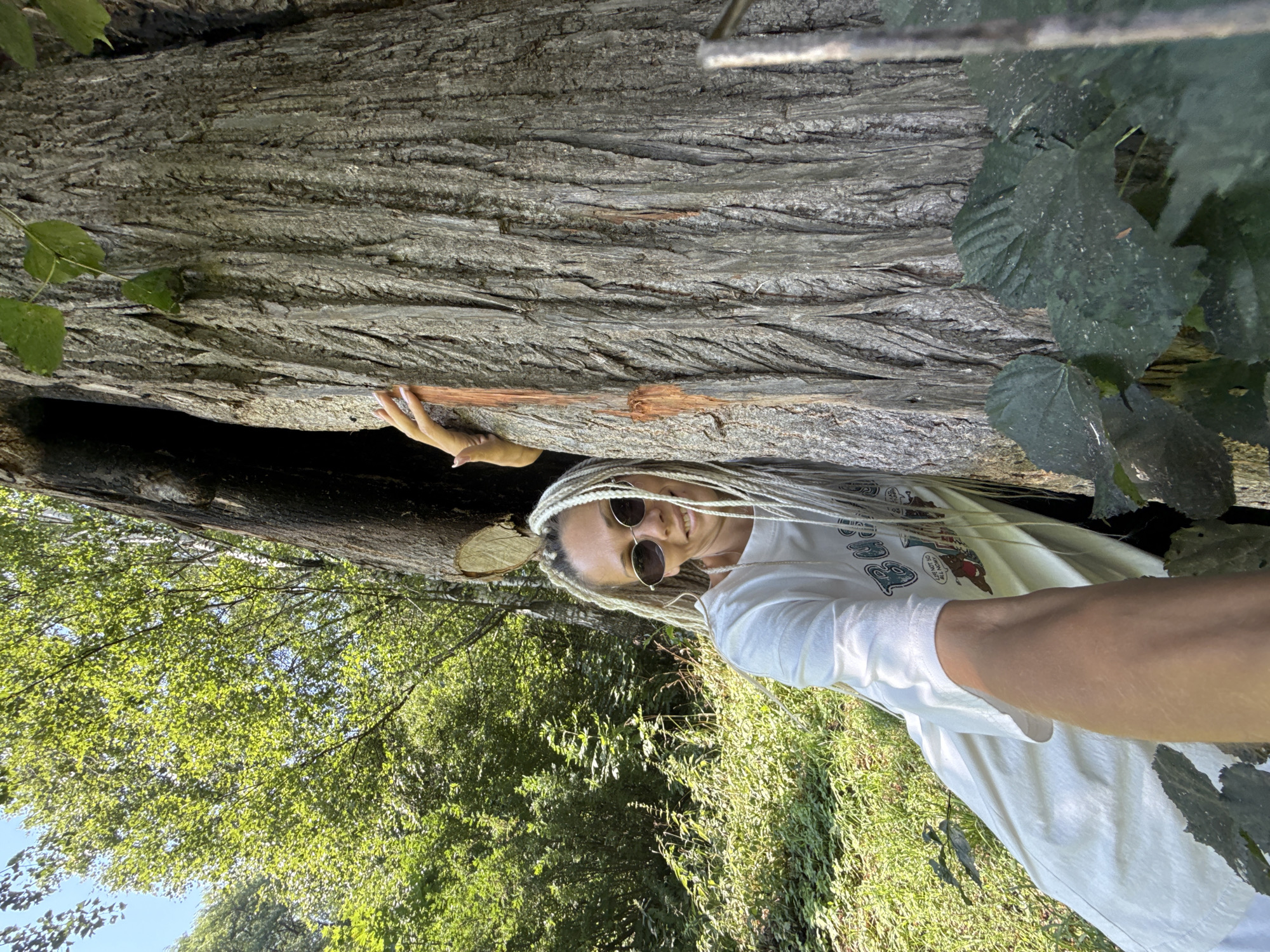 A woman with long, blonde braided hair and sunglasses is leaning against a large, rough-barked tree in a wooded area. She is wearing a white t-shirt and smiling at the camera. Her left hand is visible, resting on the tree trunk. The background shows dense green foliage and sunlight filtering through the trees. The scene appears to be outdoors, in a natural environment.